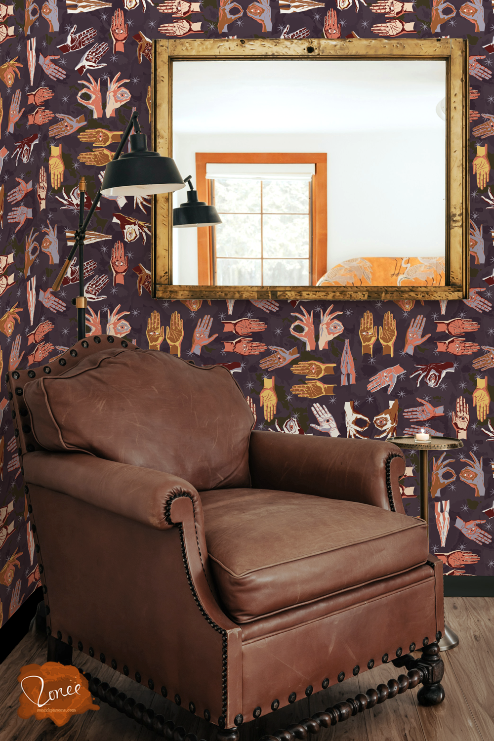 Old leather chair with a black floor lamp and vintage mirror in a room with gothic maximalist witchy wallpaper with spooky hands with eyes in purple and pink and orange.
