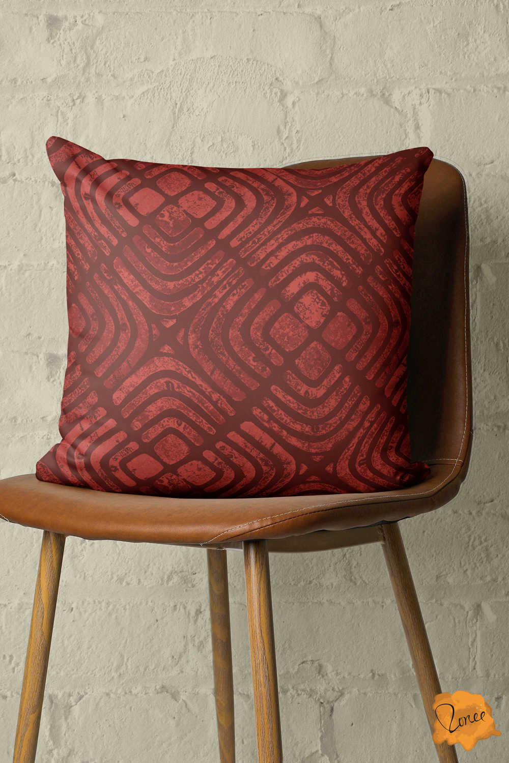 Block printed boho geometric throw pillow in deep cherry red and sepia brown on a wood and leather chair in front of a painted brick wall.
