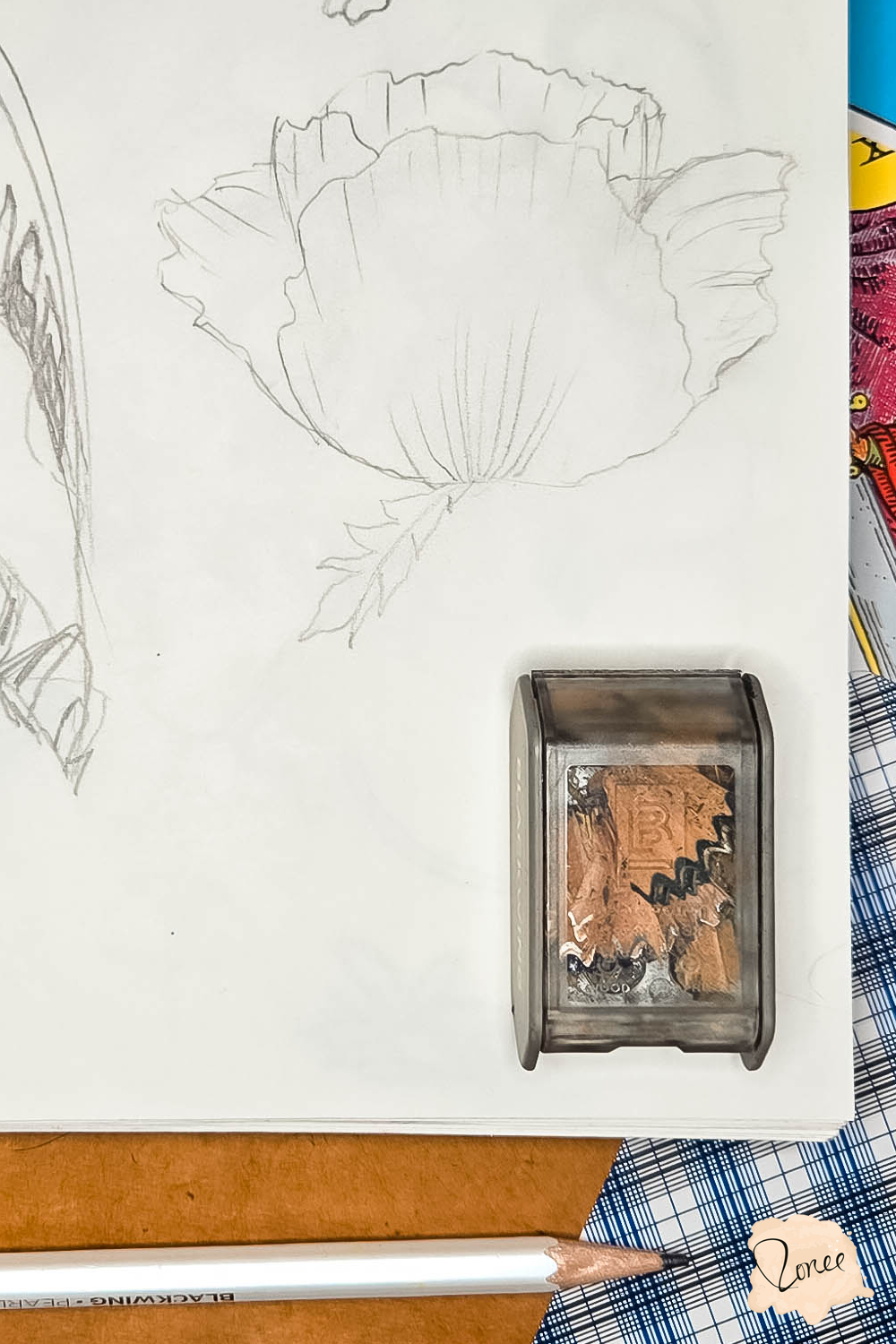 Close up flat lay image of a sketchbook with a poppy drawn roughly in pencil, a pencil sharpener with shavings is in the bottom right, a white pencil is resting along the bottom and tarot cards can be seen under the sketchbook to the right.