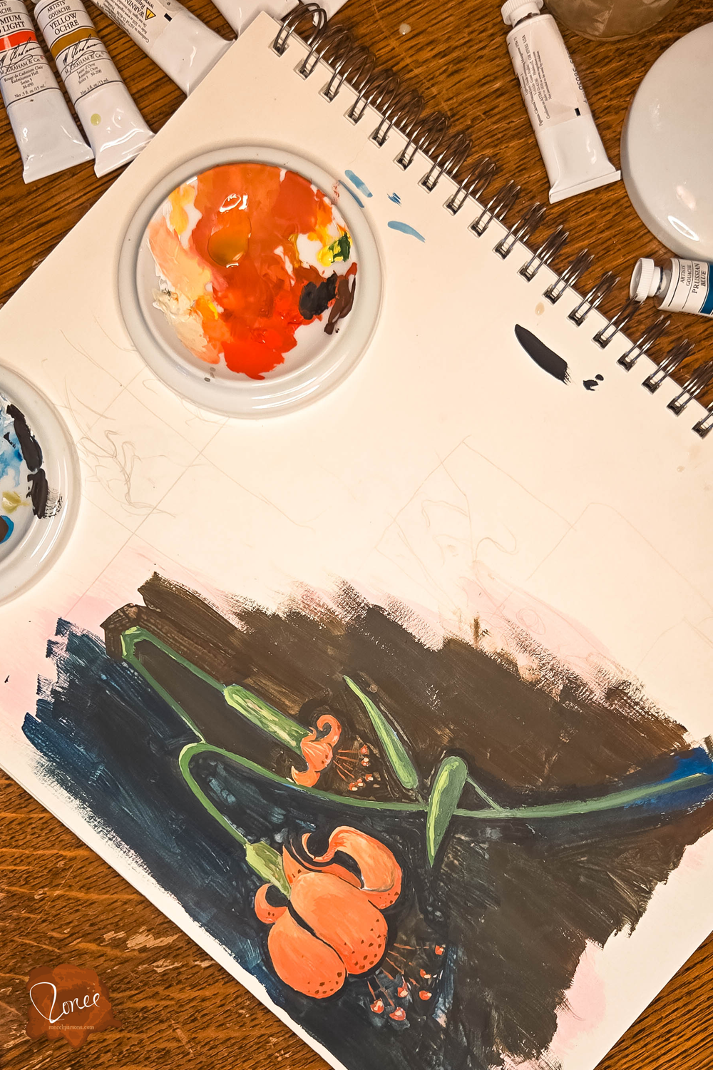 Close up flat lay image of a sketchbook with a orange gouache lily painting with paint tubes and ceramic palettes above.