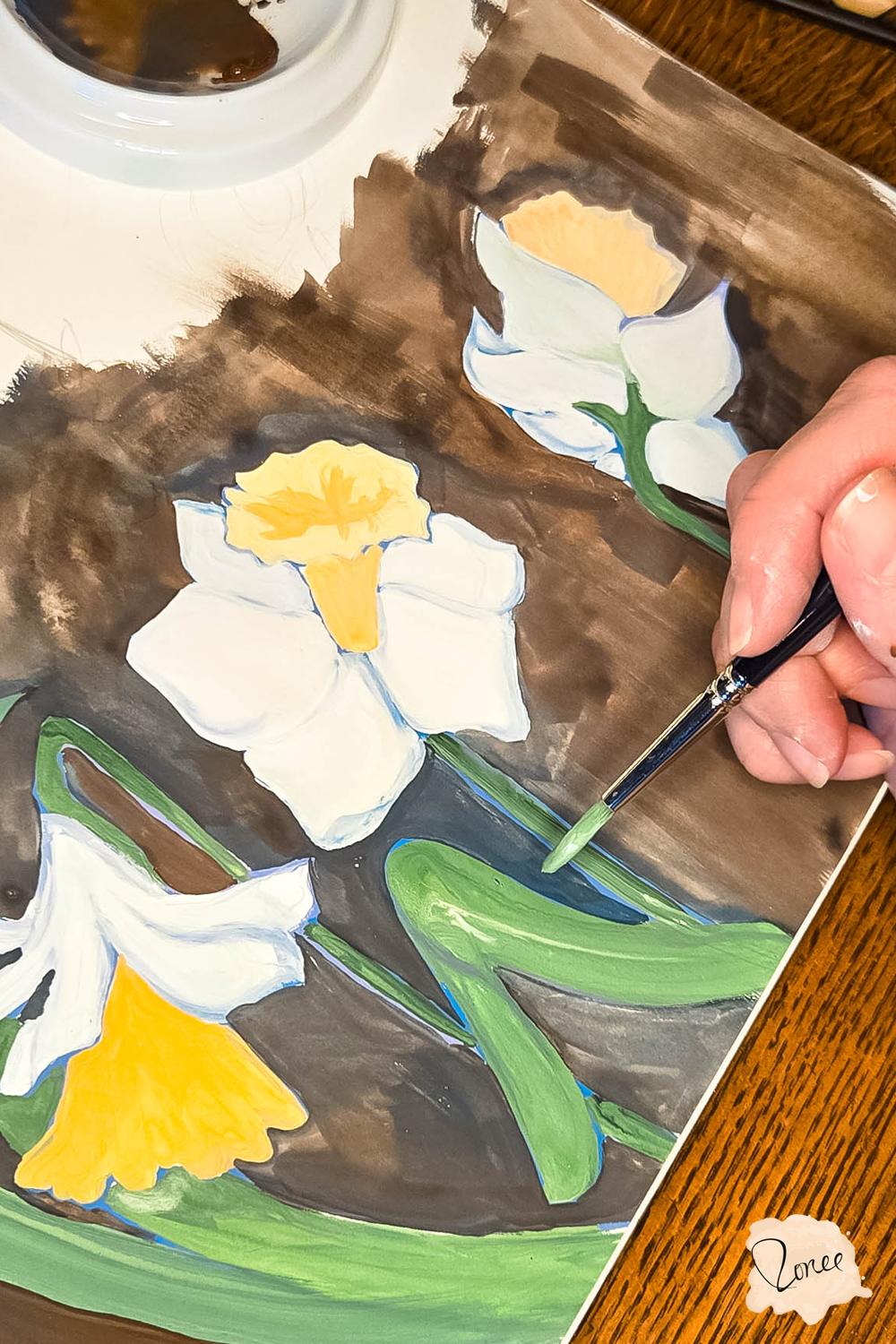 Close up flat lay image of a sketchbook with a hand painting a green daffodil leaf in gouache on series of painted daffodils..