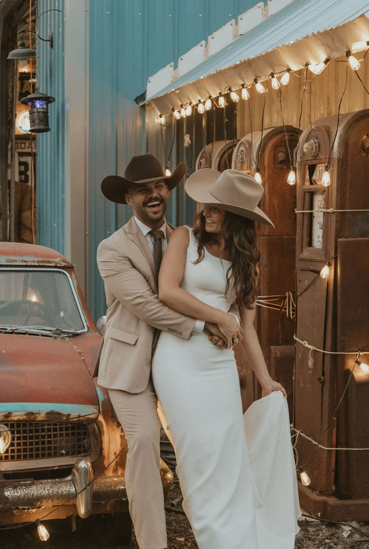 Couple in western wear posing by an antique car and string lights at Sparks Museum and Event Center