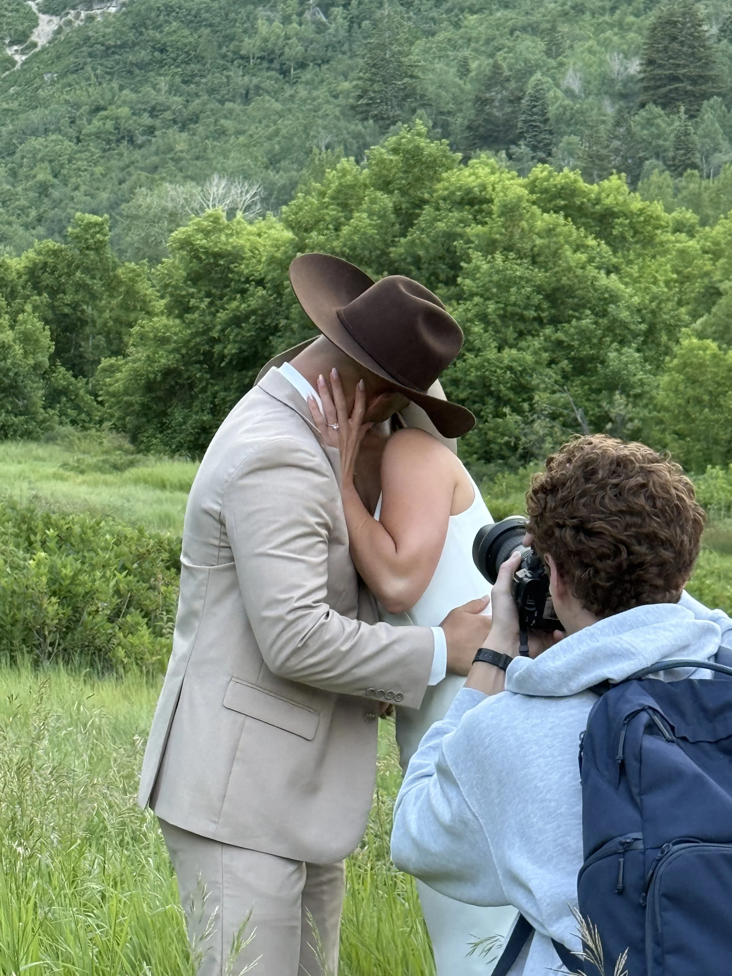Photographer capturing a tender kiss between a bride and groom in a scenic green meadow on their wedding day, with the groom wearing a tan suit and cowboy hat.
