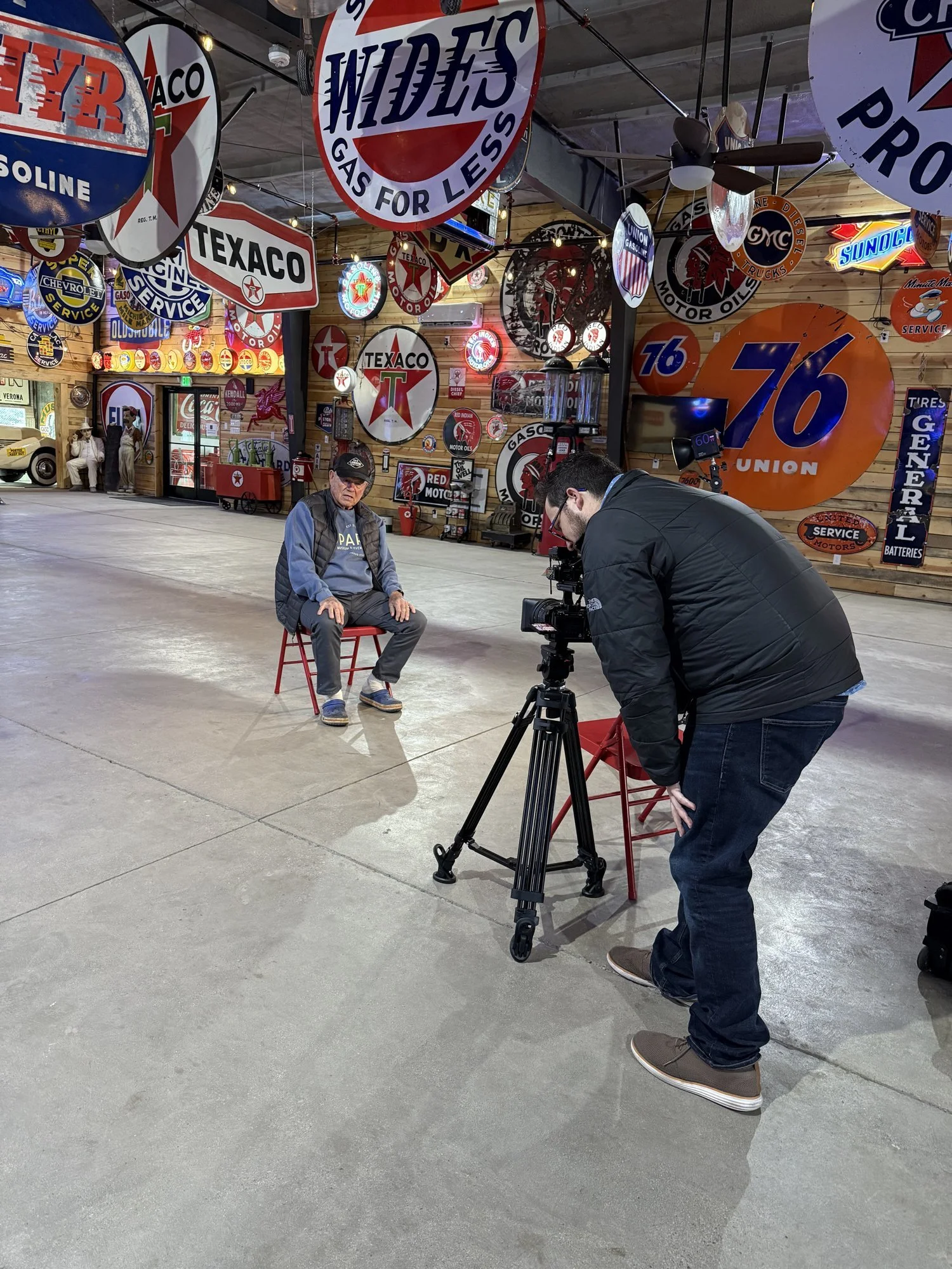 Camera operator filming an interview beside a wall of vintage gas signs at Sparks Museum and Event Center