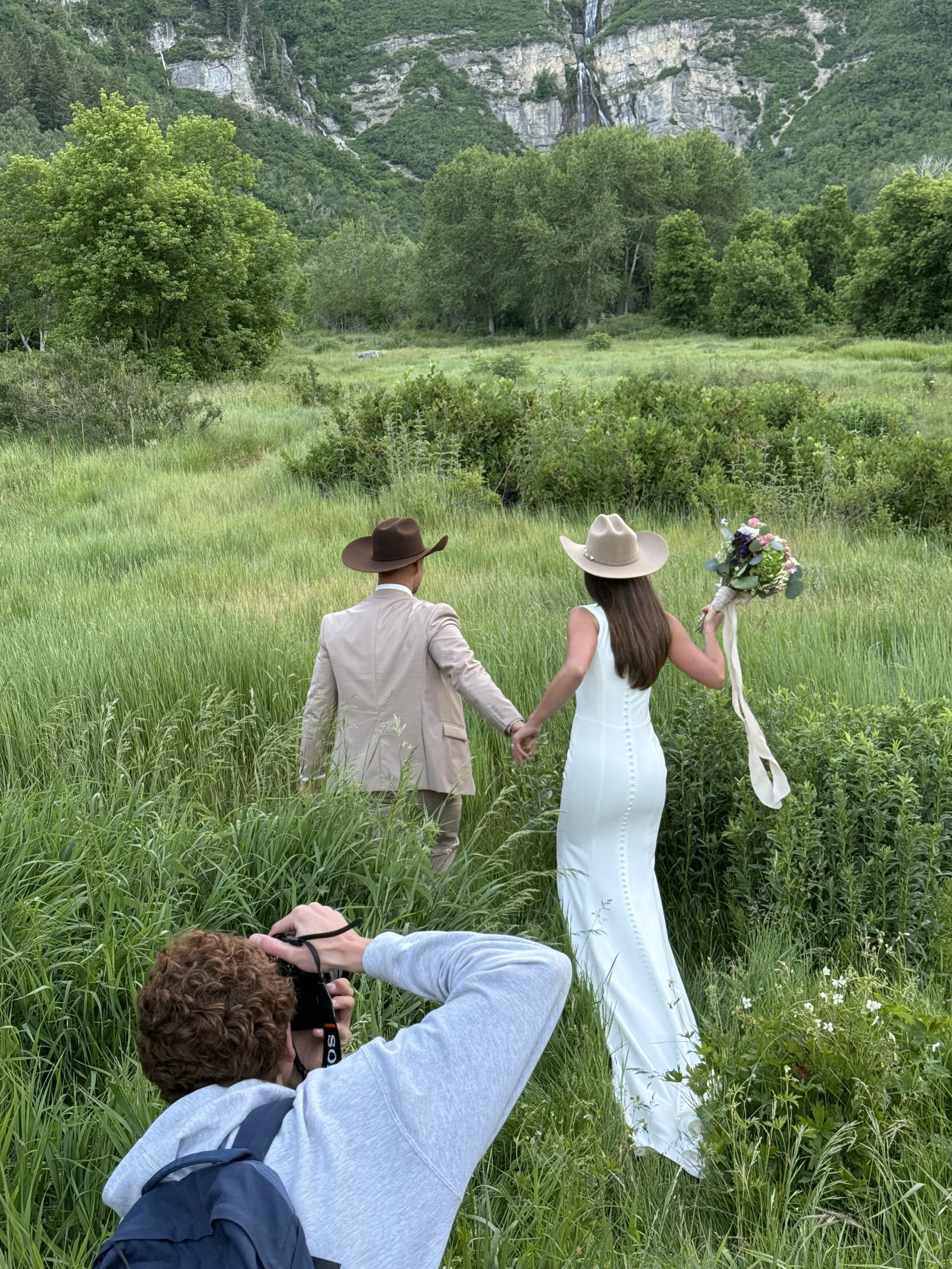 Photographer in action capturing a bride and groom walking hand-in-hand through a lush green field, with the couple dressed in western wedding attire and the photographer crouched in the grass to get the perfect shot.
