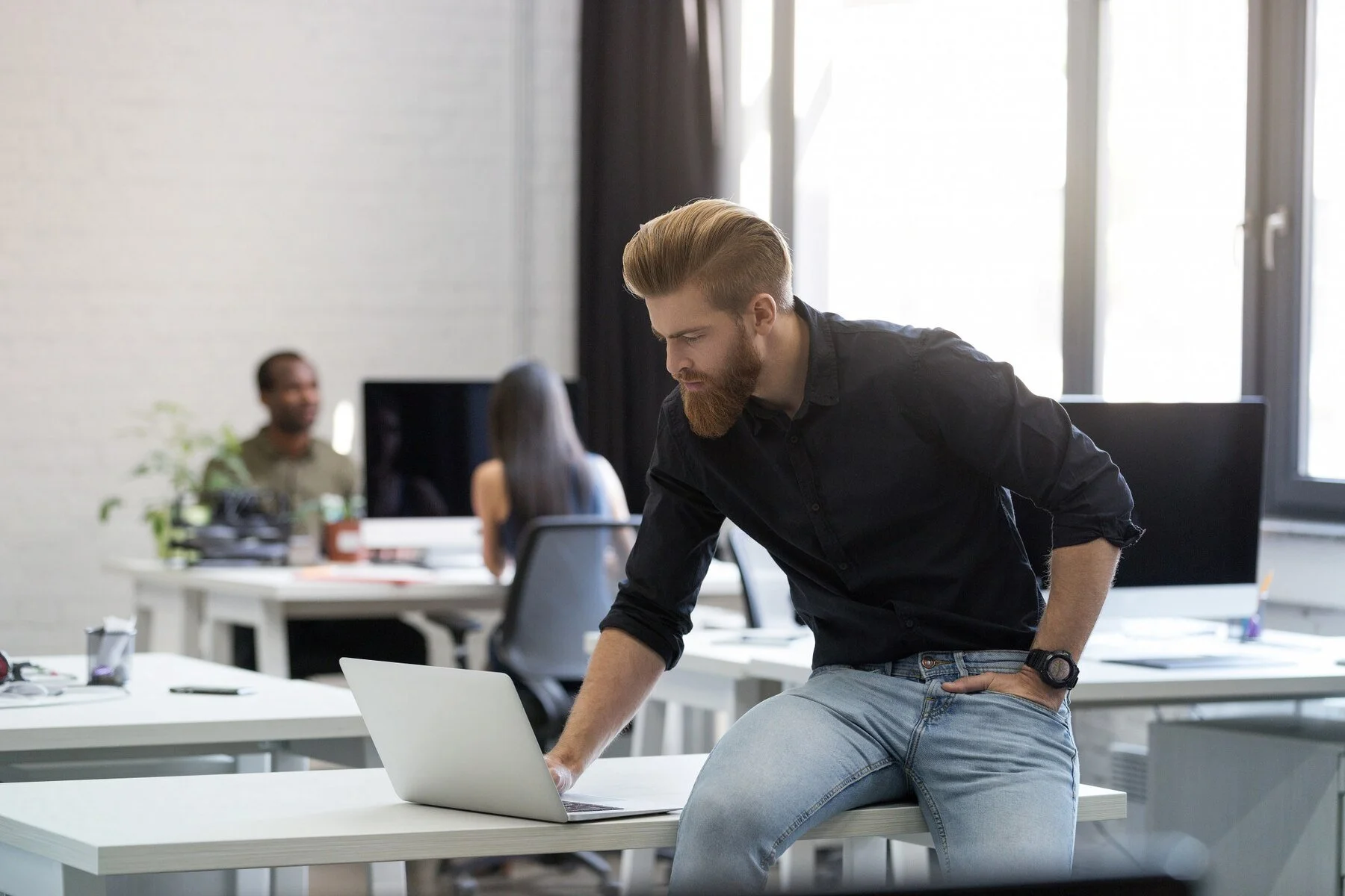 Young man with a beard in a black shirt and jeans leaning on a white table with a laptop in a bright office, with two colleagues working in the background – representing the Wiseep team