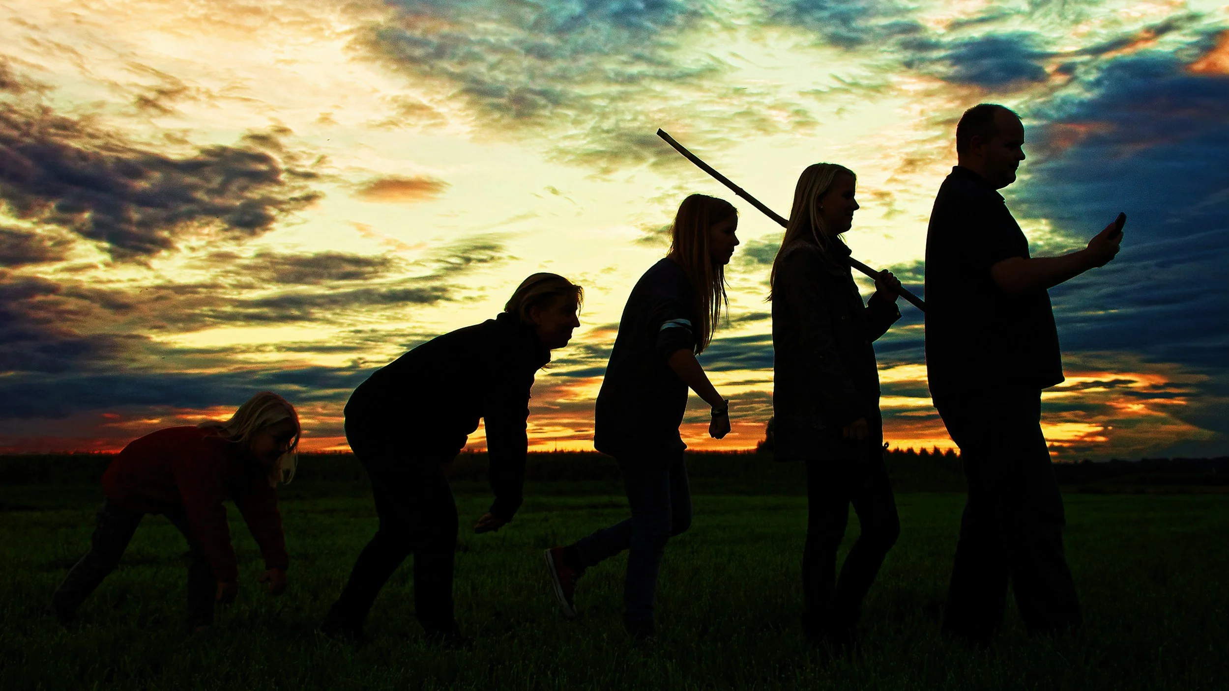 Silhouettes of five people standing in a field during sunset, with one person holding a sword and another looking at a smartphone.