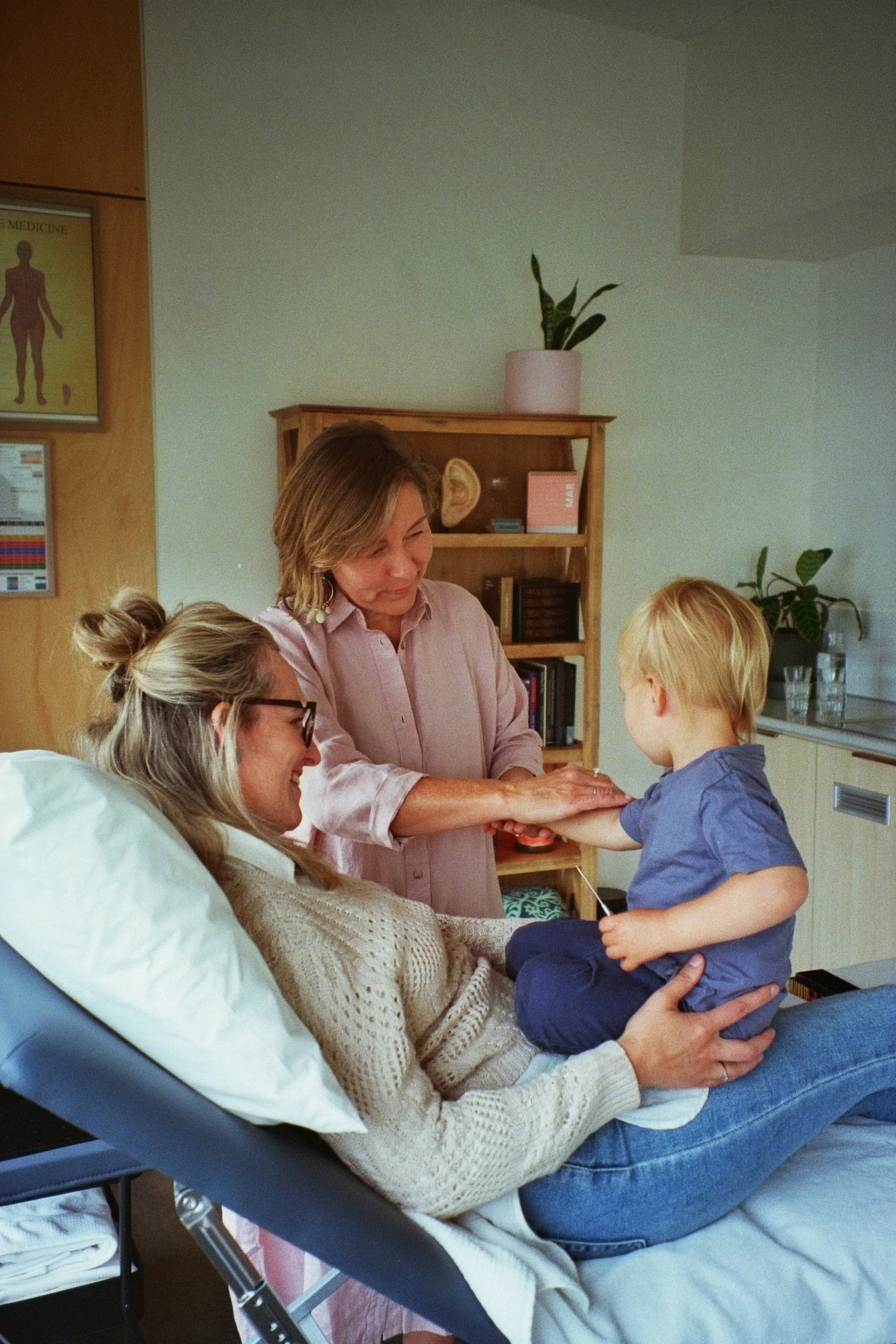 A woman lying on a hospital bed with an IV, another woman sitting beside her, and a young boy standing in front of the woman, holding her hand in a comforting manner.
