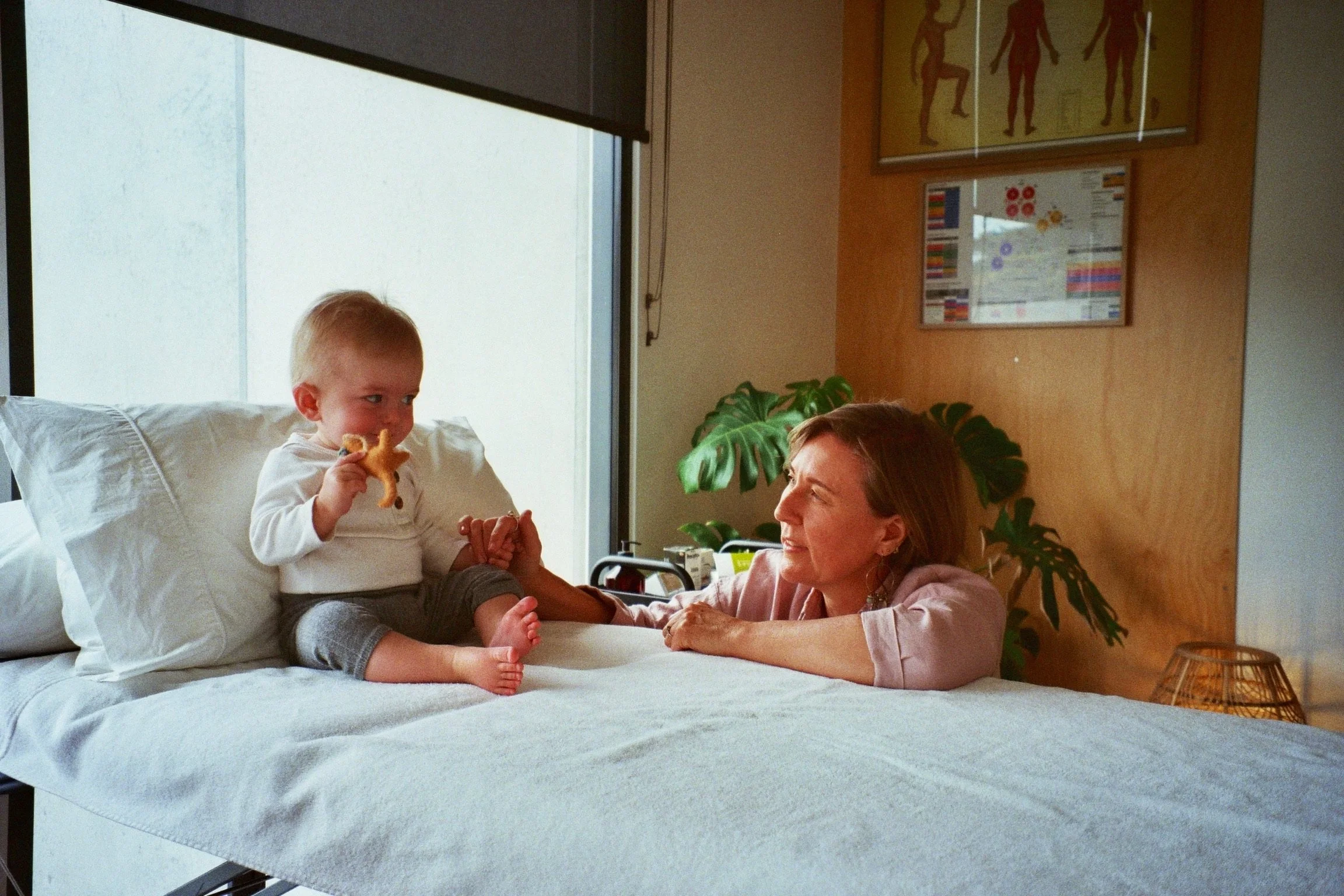 A young boy sitting on a hospital bed holding a stuffed animal, smiling and looking at a woman lying in the bed with her head propped up on pillows. The woman is smiling back at the boy. There are medical equipment and plants in the background.