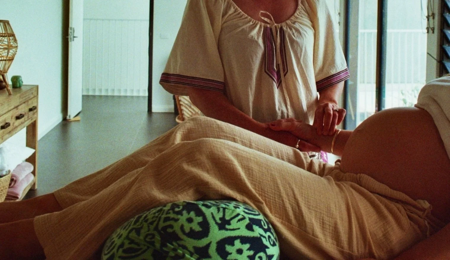 A person receiving a massage from a massage therapist in a room with natural lighting, tiled floors, and sliding glass doors.