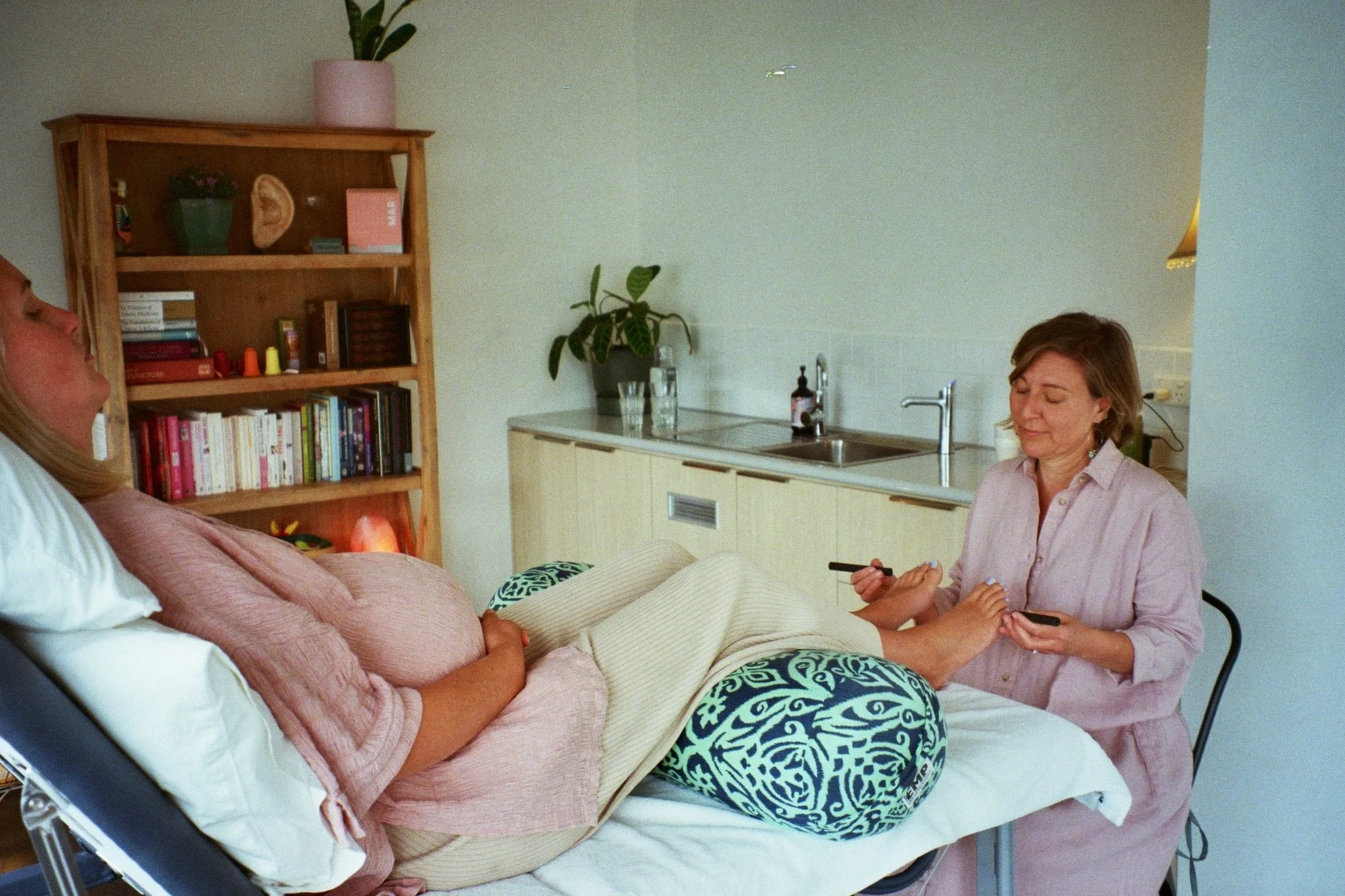 A woman lying in a hospital bed with a nurse or caregiver sitting beside her, holding her hand while she talks. The room has a bookshelf, plants, and medical supplies in the background.