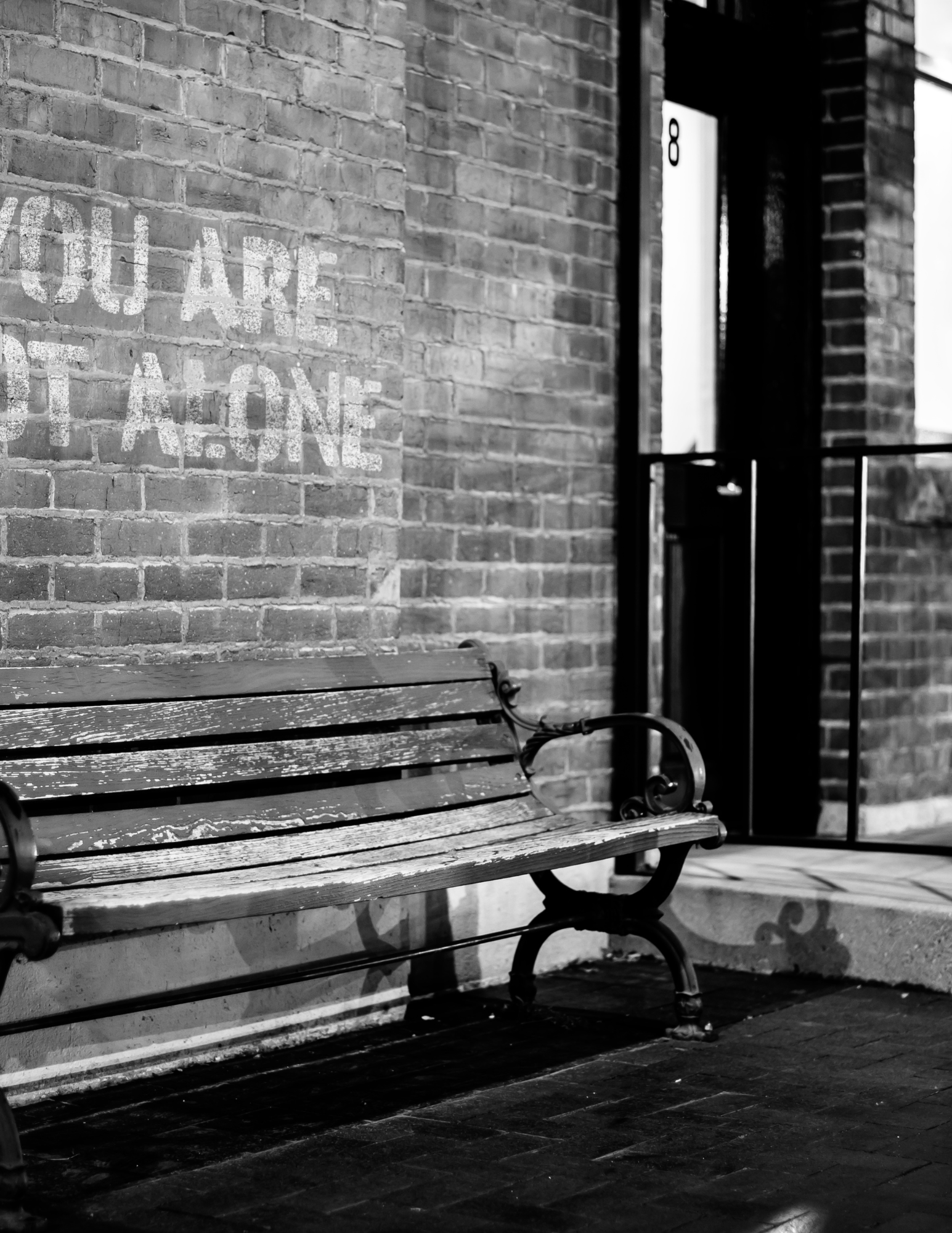 Black and white photo of an old wooden bench against a brick wall with the words "YOU ARE NOT ALONE" painted on it.