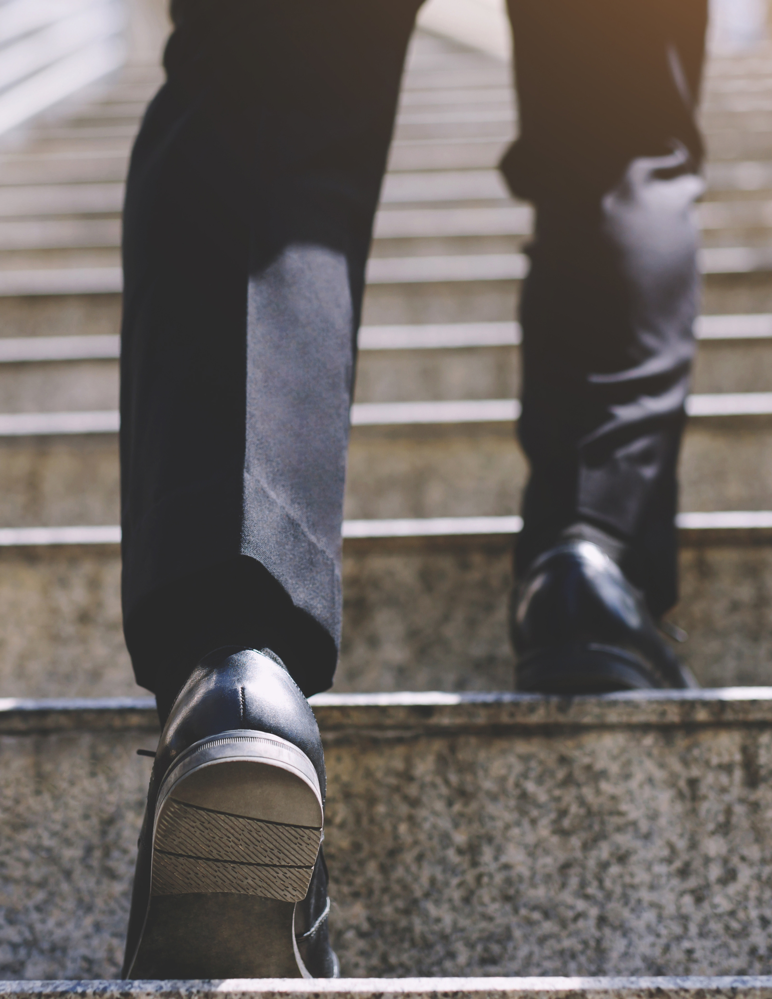 Close-up of a person in dark pants and black leather shoes walking up stone stairs.