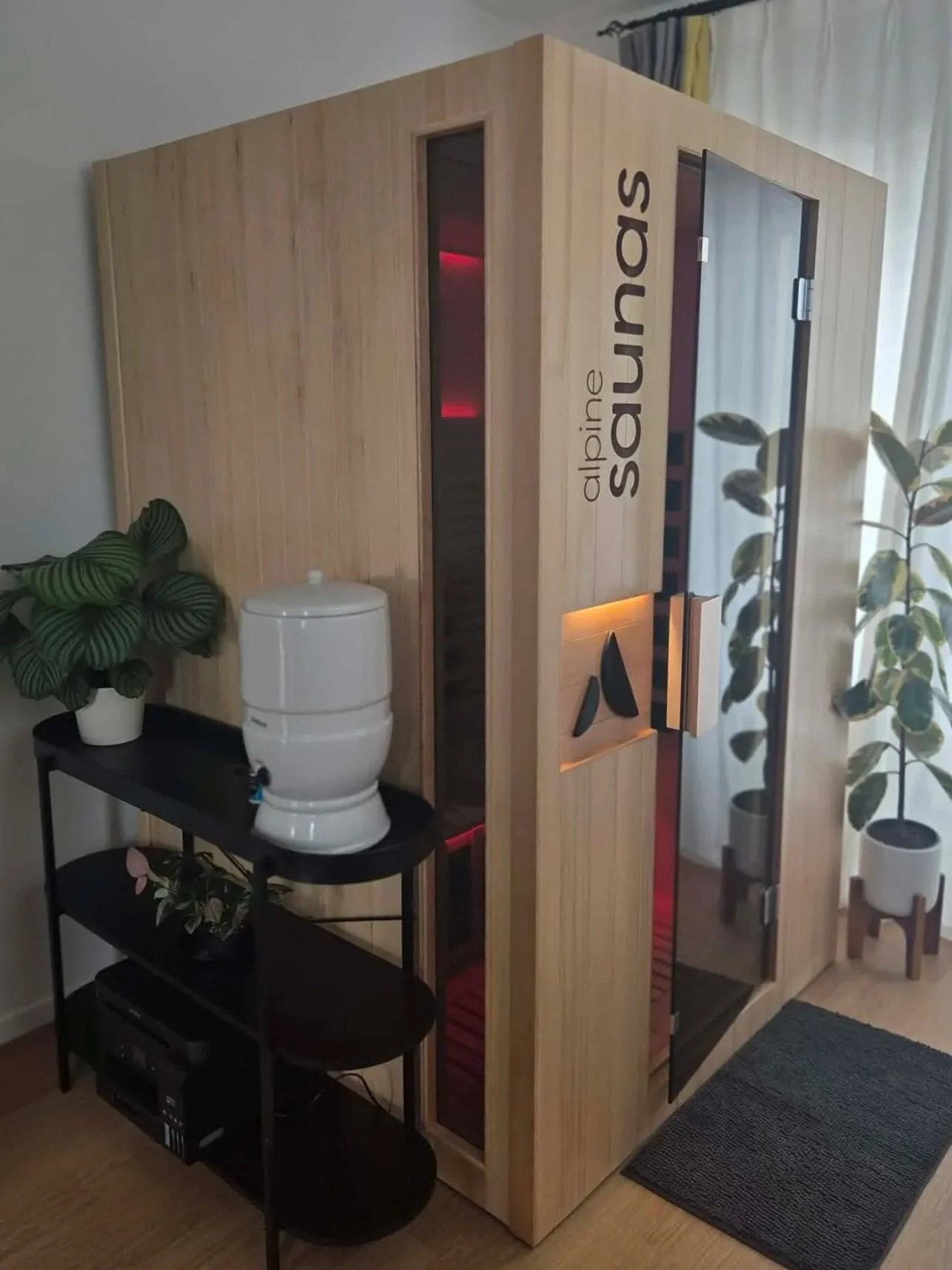 A wooden infrared sauna named 'alpine sauna' with glass doors, positioned next to houseplants and a black shelving unit with a white device on top, located in a room with curtains and hardwood flooring.