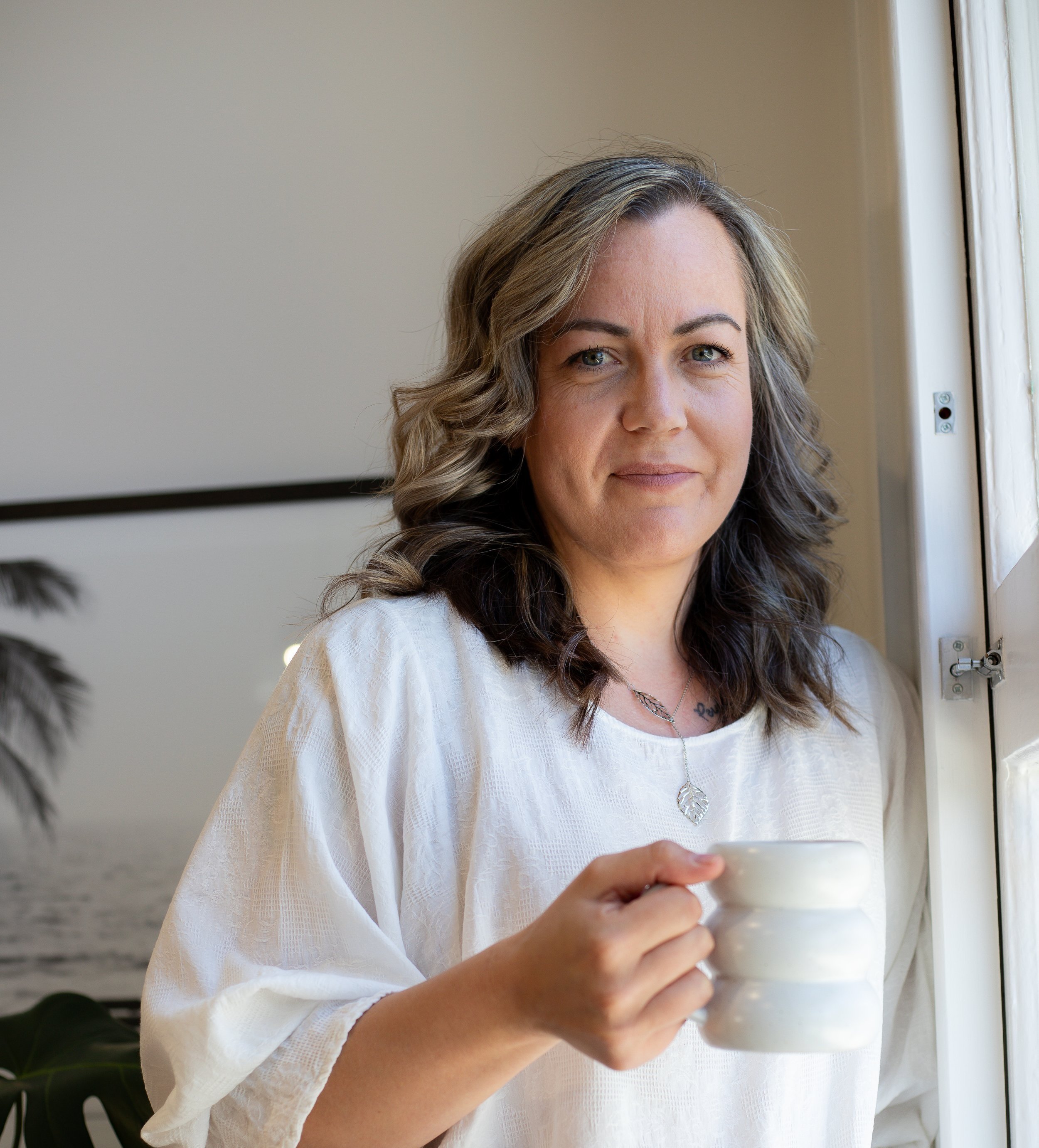 A woman with wavy hair holding a white mug, standing near a window with sunlight, in a room with minimal decor.