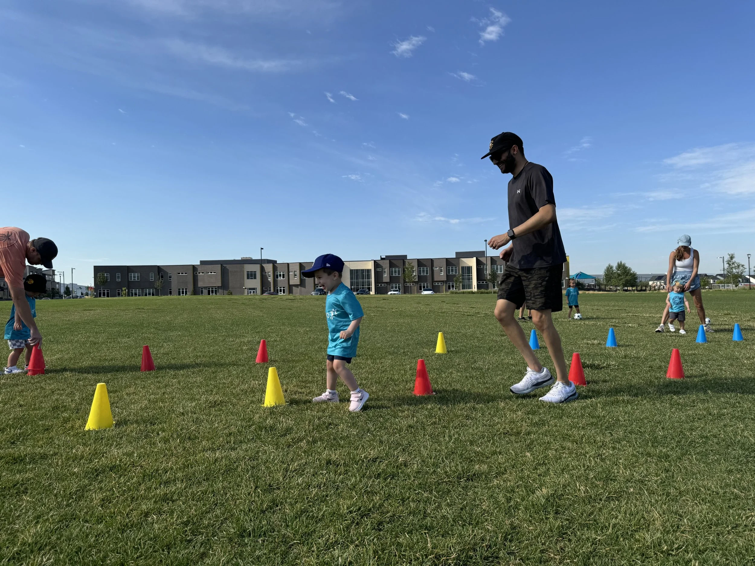 Adults and children participating in an outdoor activity on a grassy field, with colorful cones set up for a race or obstacle course, under a partly cloudy blue sky.