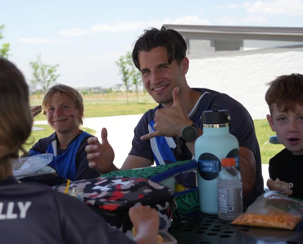 A man and two boys sitting at a table outdoors, smiling and making a thumbs-up gesture, with sports water bottles and snacks on the table.