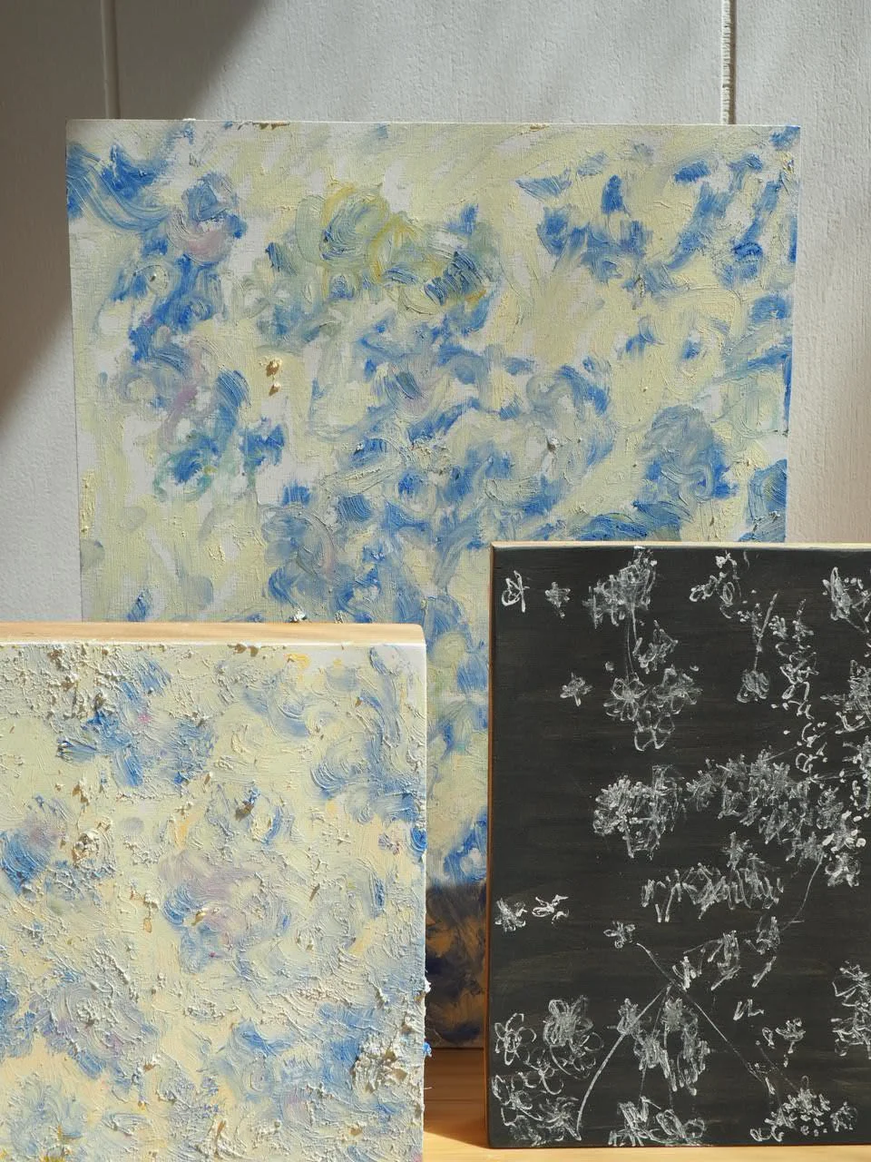 Three abstract painter's canvases, one with blue and cream swirling strokes, another with textured cream and blue, and a third black canvas with white floral sketches.