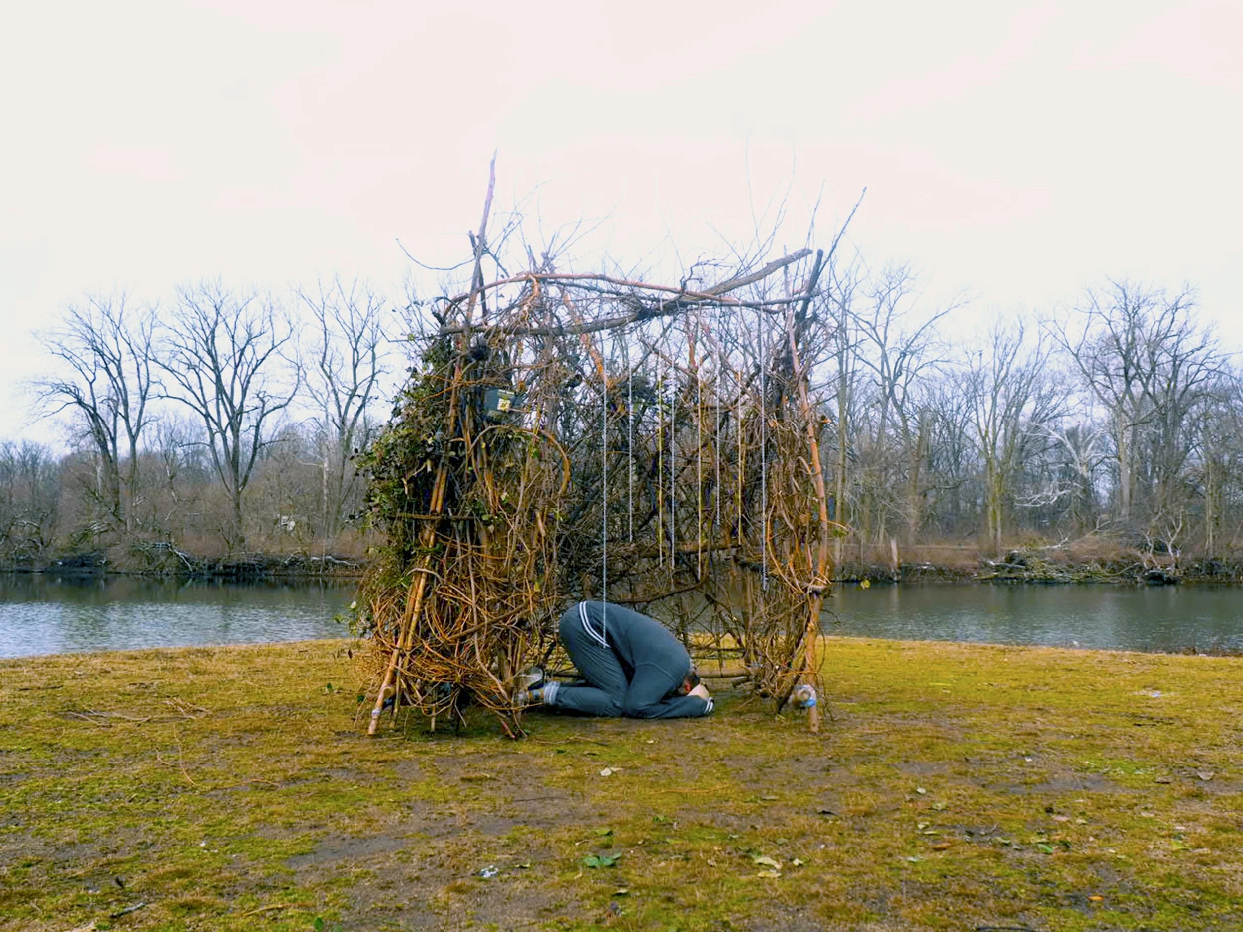 Person kneeling and working on a small man-made structure made of sticks and branches near a body of water, surrounded by leafless trees.