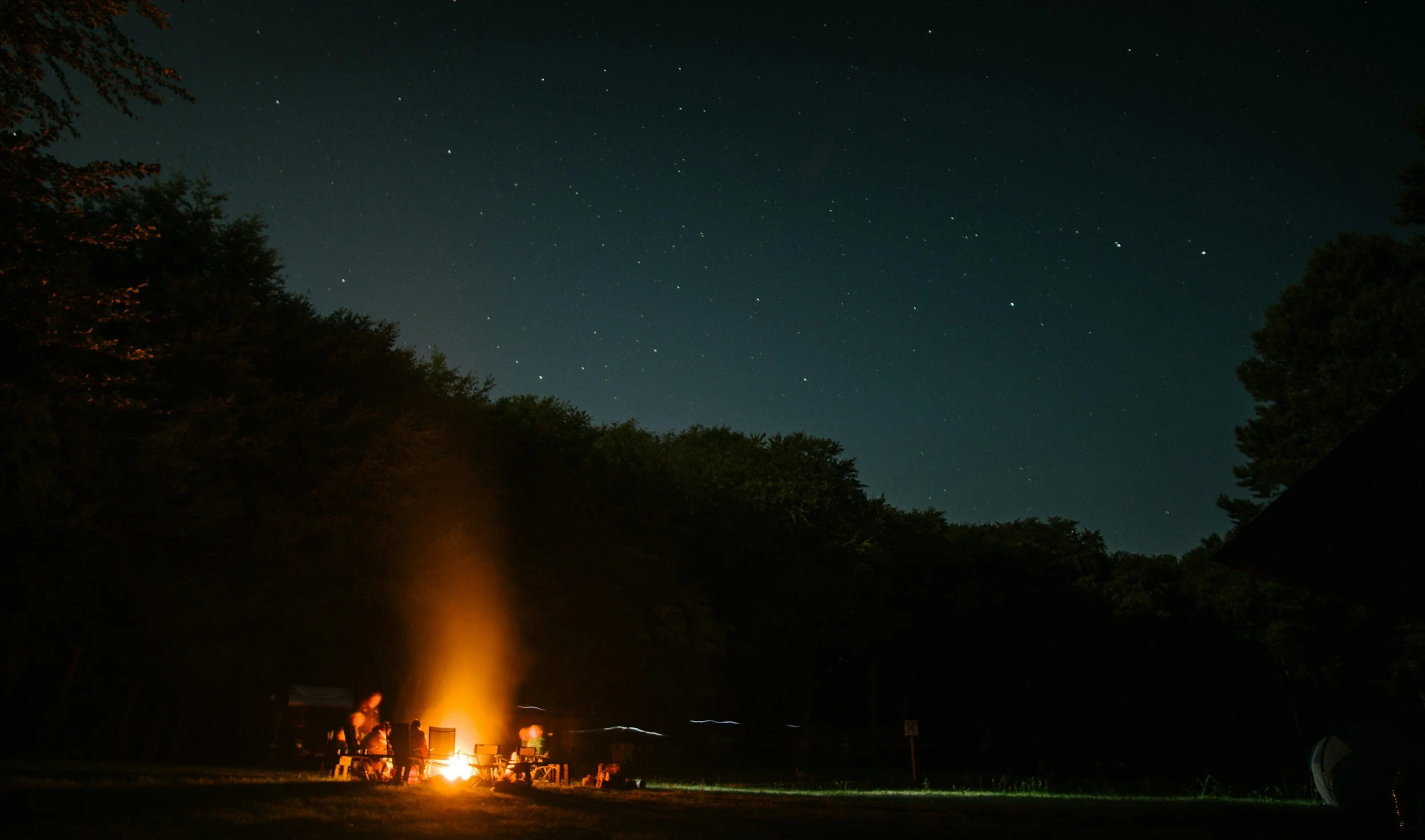 people sitting around a fire at night with a starry sky above and trees and hills silhouetted against the sky