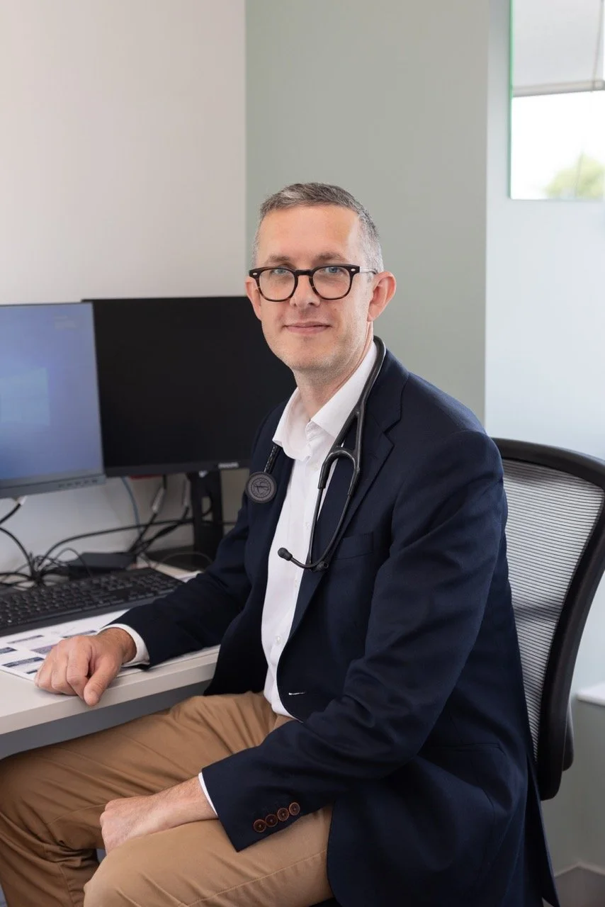 Dr Daniel Garner sitting at a desk in an office, wearing glasses, a white shirt, a dark blazer, and a stethoscope around his neck.