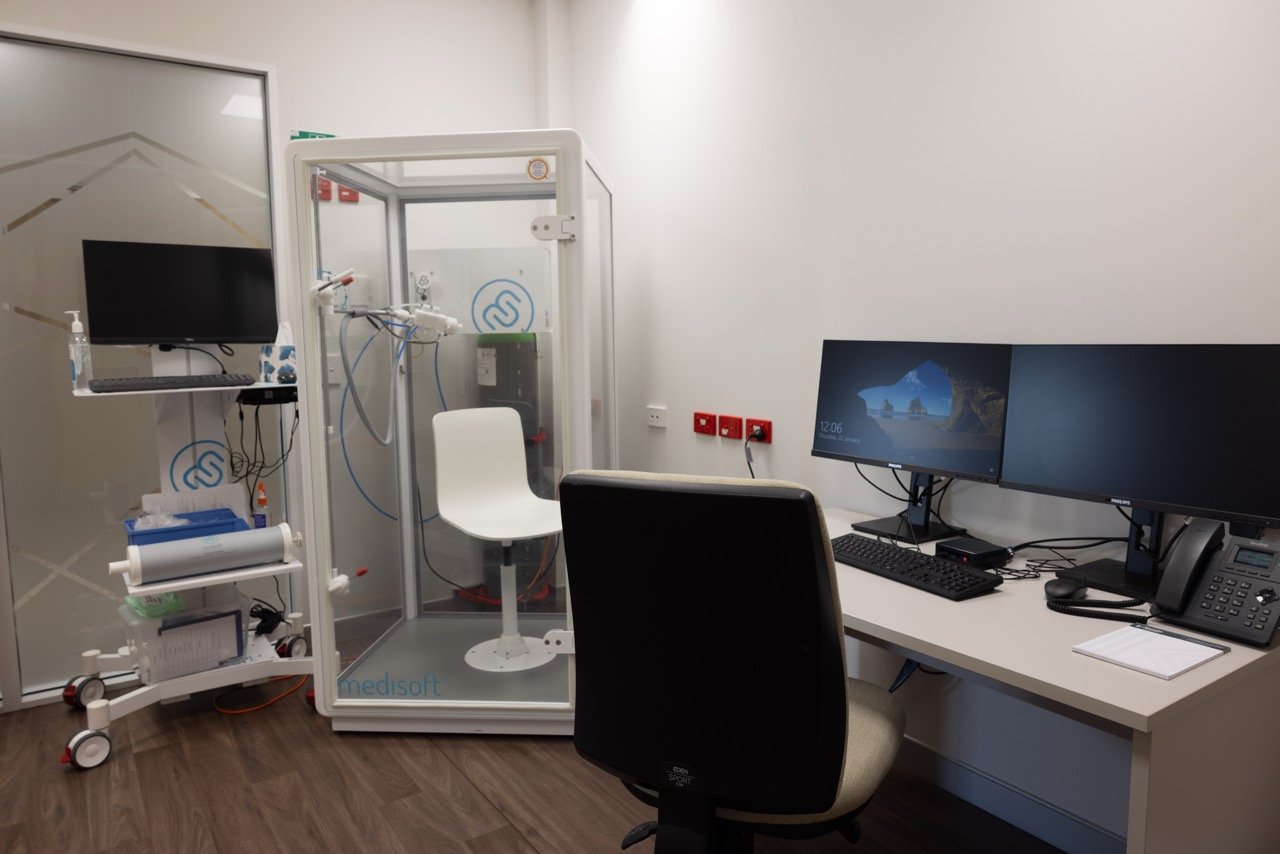 Hawke's Bay Respiratory and Sleep medical room with a white chair inside a transparent enclosure, two computer monitors, a computer keyboard, telephone, and notepad on a white desk.