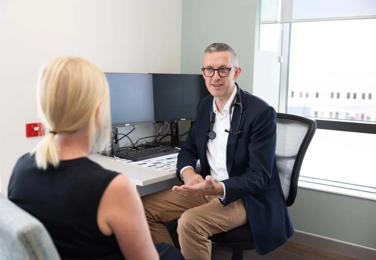 Dr Daniel Garner wearing glasses, a stethoscope around his neck, and a dark blazer, sitting and talking to a blonde woman with her hair in a ponytail, in an office with a desk, multiple computer monitors, and a large window showing building views.