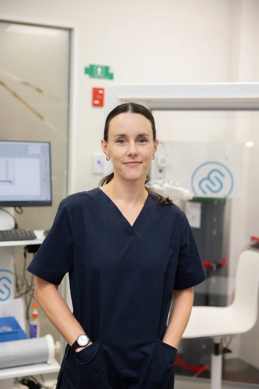 Resipratory and Sleep Physiologist Jemma Robyns in navy scrubs standing in a medical laboratory or clinic, with medical equipment and a computer in the background.