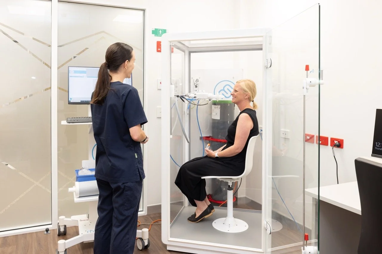 A woman participating in a medical or scientific test inside a sealed transparent chamber with Respiratory and Sleep Physiologist Jemma Robyns observing.