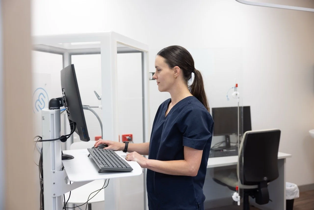 Respiratory and Sleep Physiologist Jemma Robyns in navy scrubs working on a computer in a lab or medical office with medical equipment and monitor in the background.