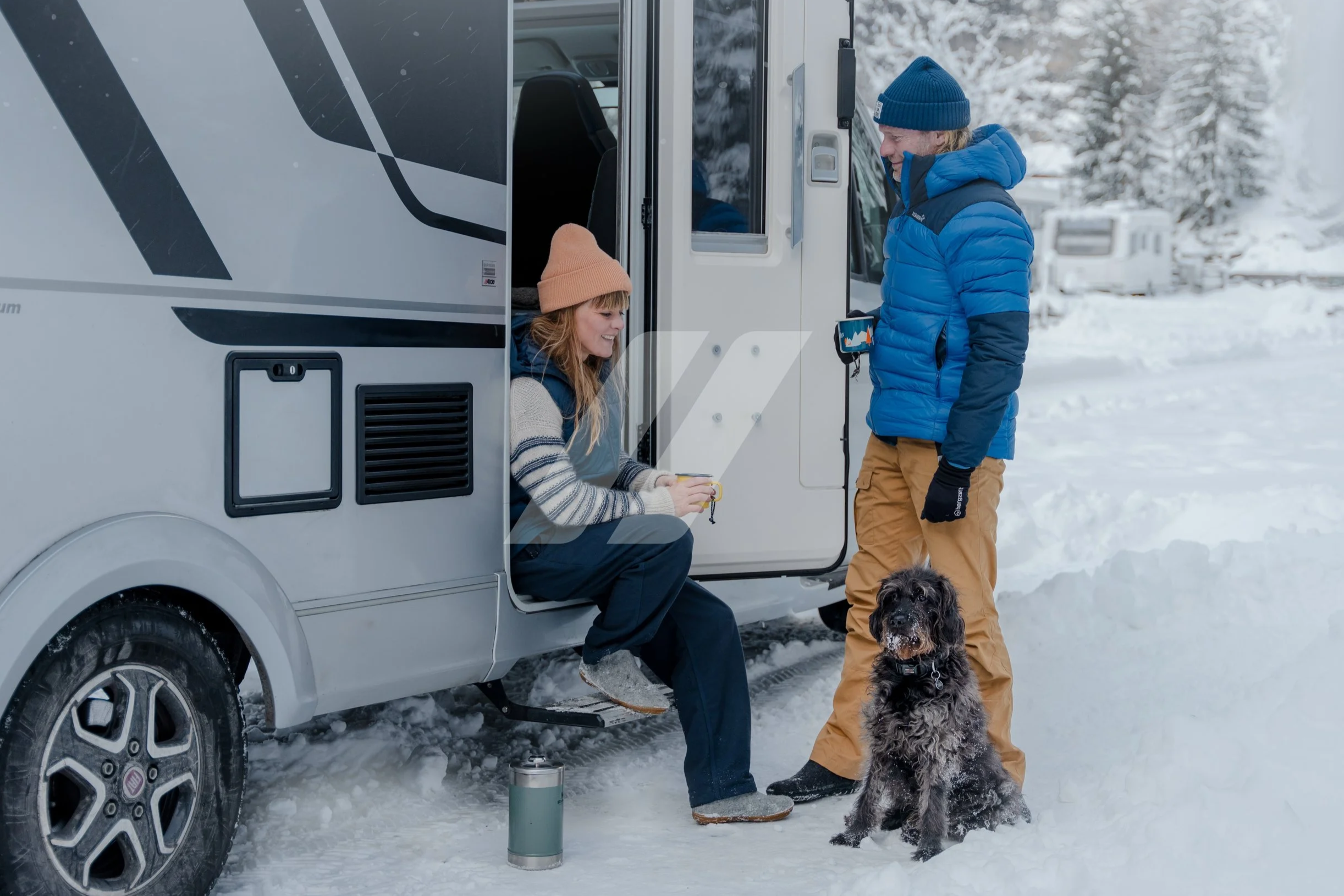 A woman with red hair sitting on the step of a white camper van, wearing a beige beanie and a striped sweater, smiling at a man in a blue puffy jacket and tan pants, who is holding a leash with a black and white dog. The man is wearing a blue beanie.