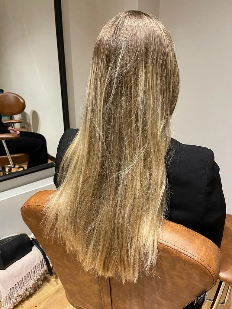 Woman with long blonde hair sitting on a brown chair, viewed from the back, inside a salon.