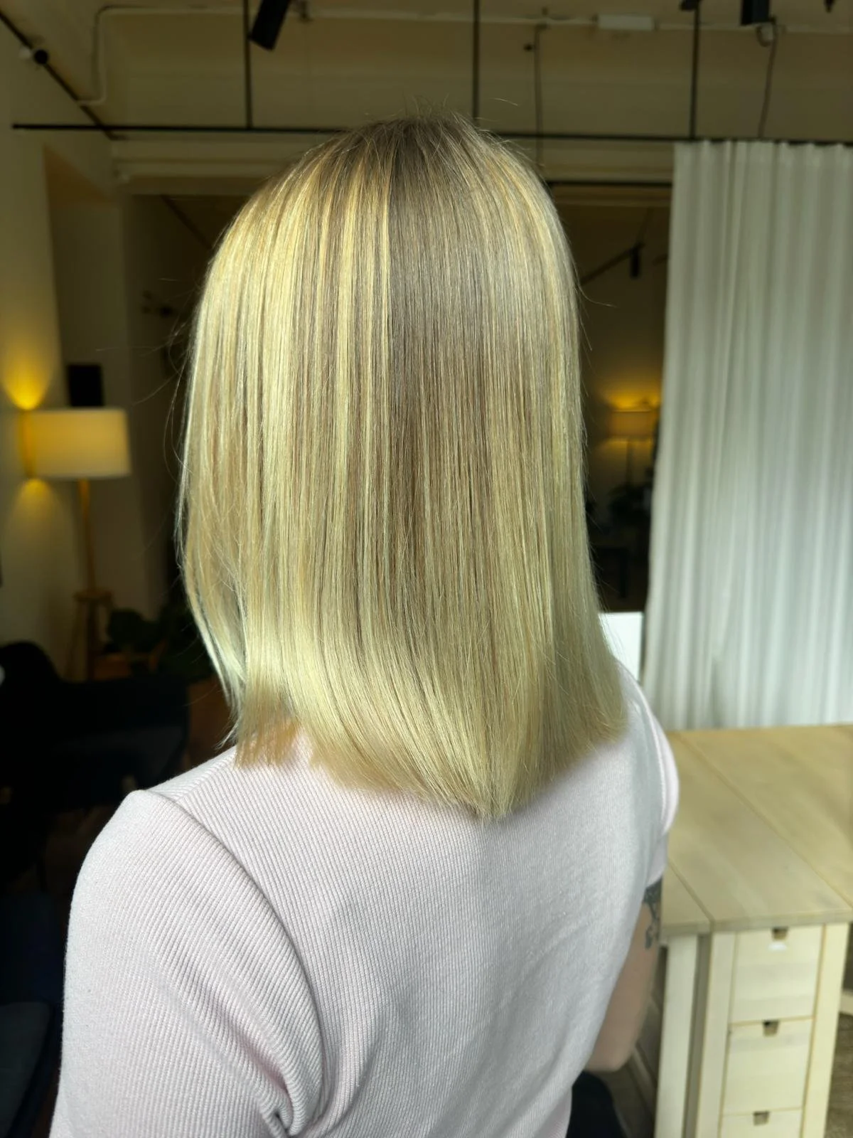 Person with straight, shoulder-length blonde hair wearing a light-colored top, seen from the back, in a room with a lamp and white curtain.