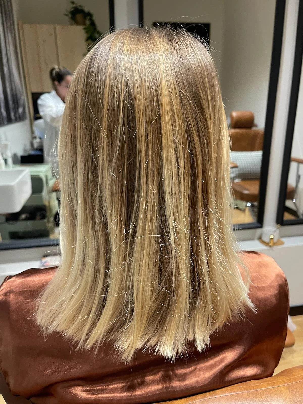 Back view of a person with shoulder-length blonde hair sitting in a salon chair.