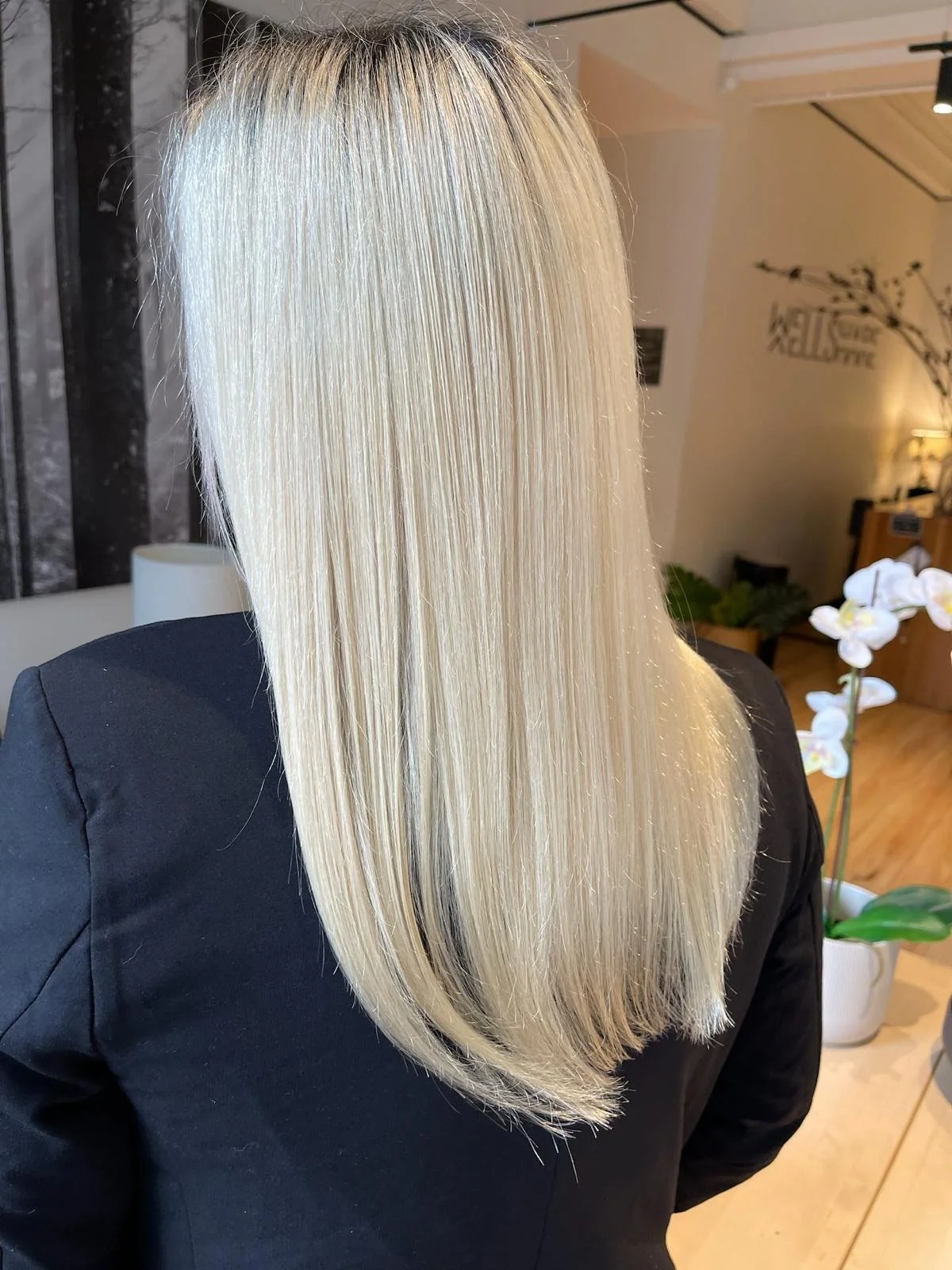 Long straight blonde hair on a person wearing a black jacket, inside a salon environment with a potted orchid and minimalist decor.
