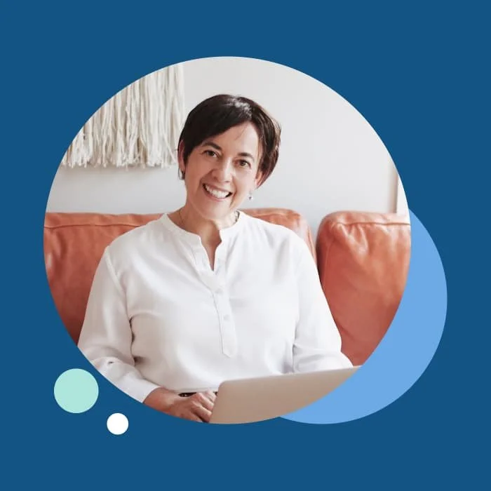 A woman with short dark hair smiling while sitting on a couch with a laptop in her lap.