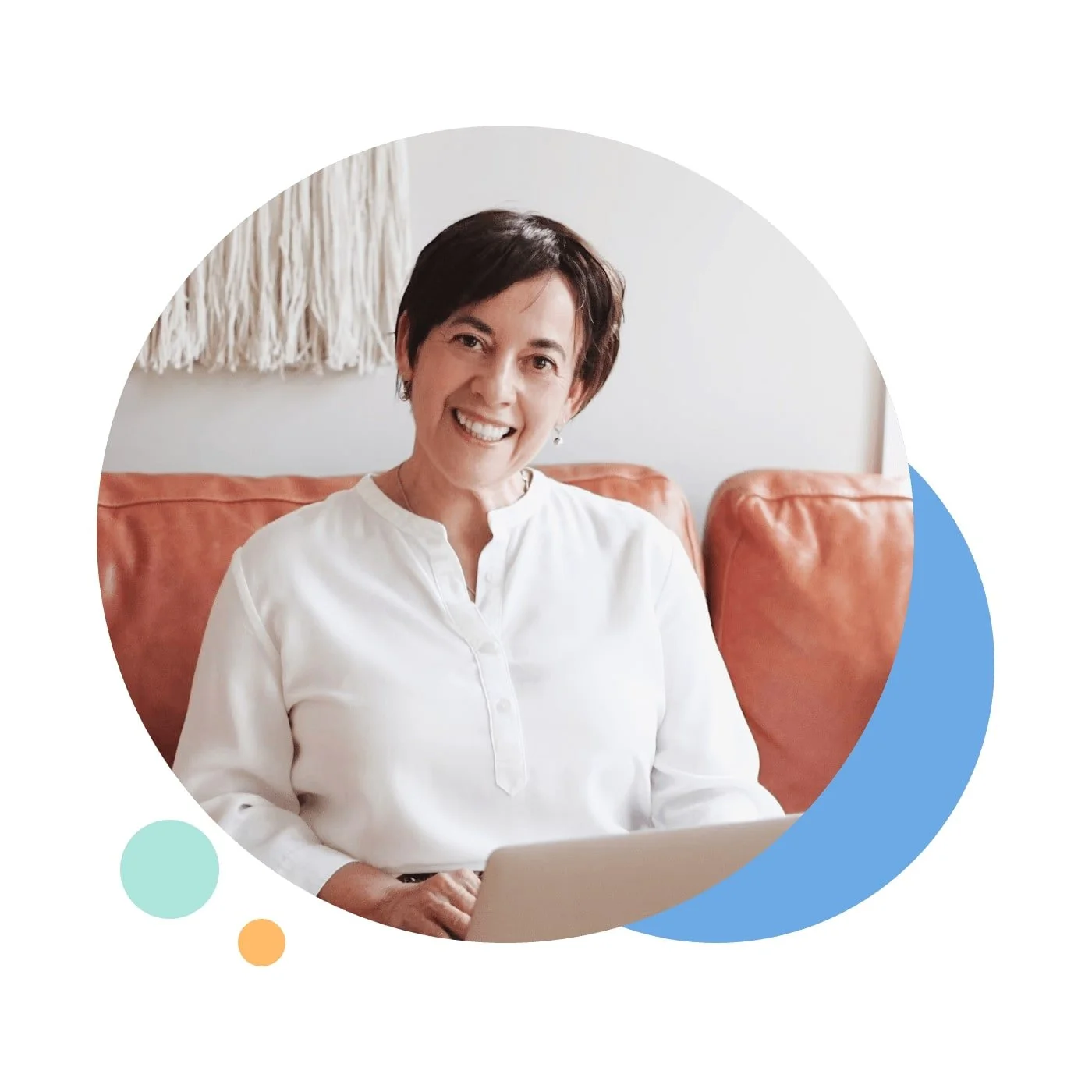 A woman smiling while sitting on a brown leather sofa, working on a laptop, with a white wall and decorative wall hanging behind her.
