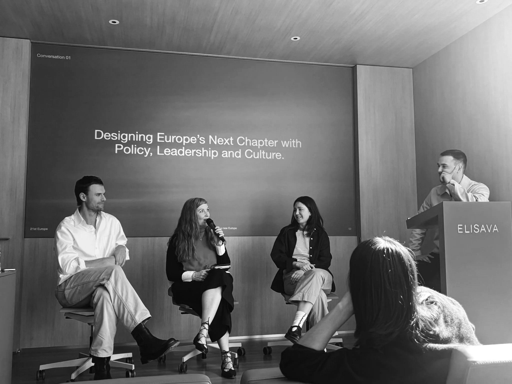 Panel discussion at a conference titled 'Designing Europe's Next Chapter with Policy, Leadership and Culture', with four individuals seated on chairs on stage. One woman is speaking into a microphone, and the others are listening. A man is standing at a podium with a name tag 'ELISAVA,' and a presentation screen behind them displays the event title.
