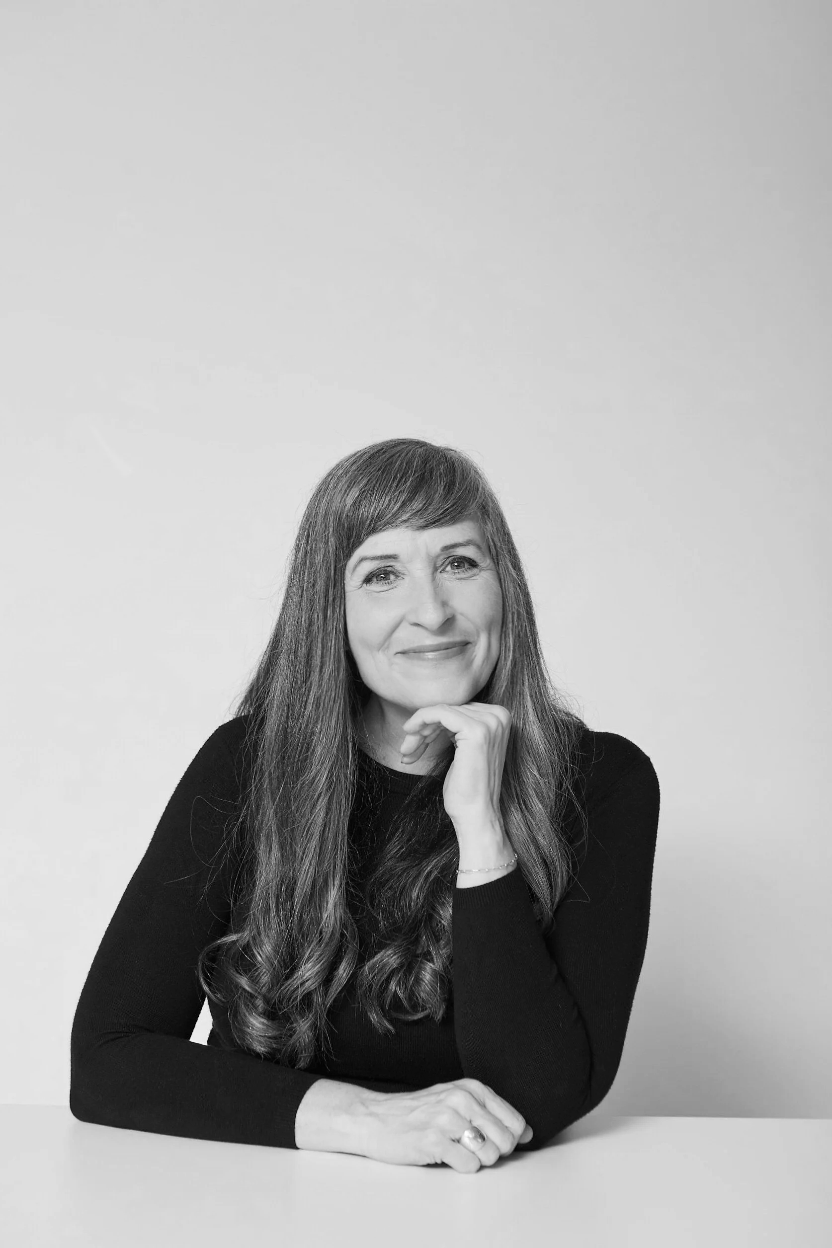 A black and white photo of a woman with long hair, sitting at a table with one hand resting under her chin, smiling softly, against a plain light background.