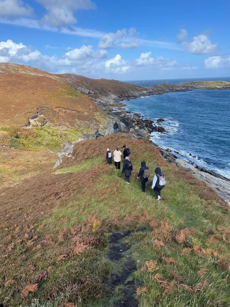 Group of people walking along a narrow coastal trail with grassy and rocky terrain, overlooking an ocean and cloudy sky.