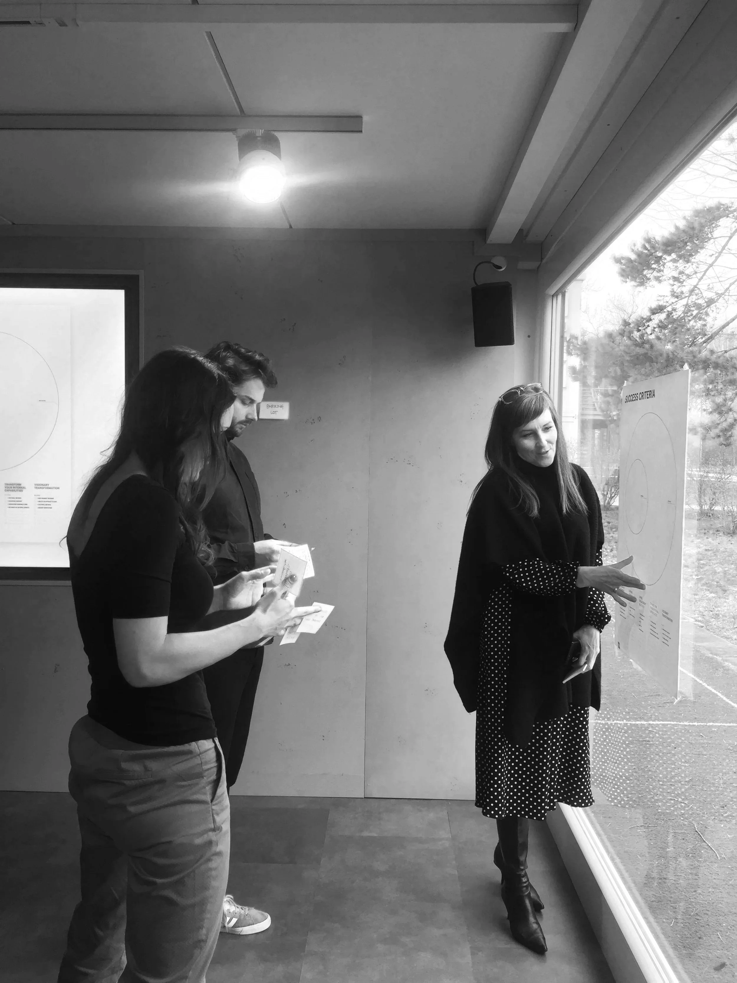 Three people inside a room near a window, with two of them reading papers and one looking at a whiteboard.