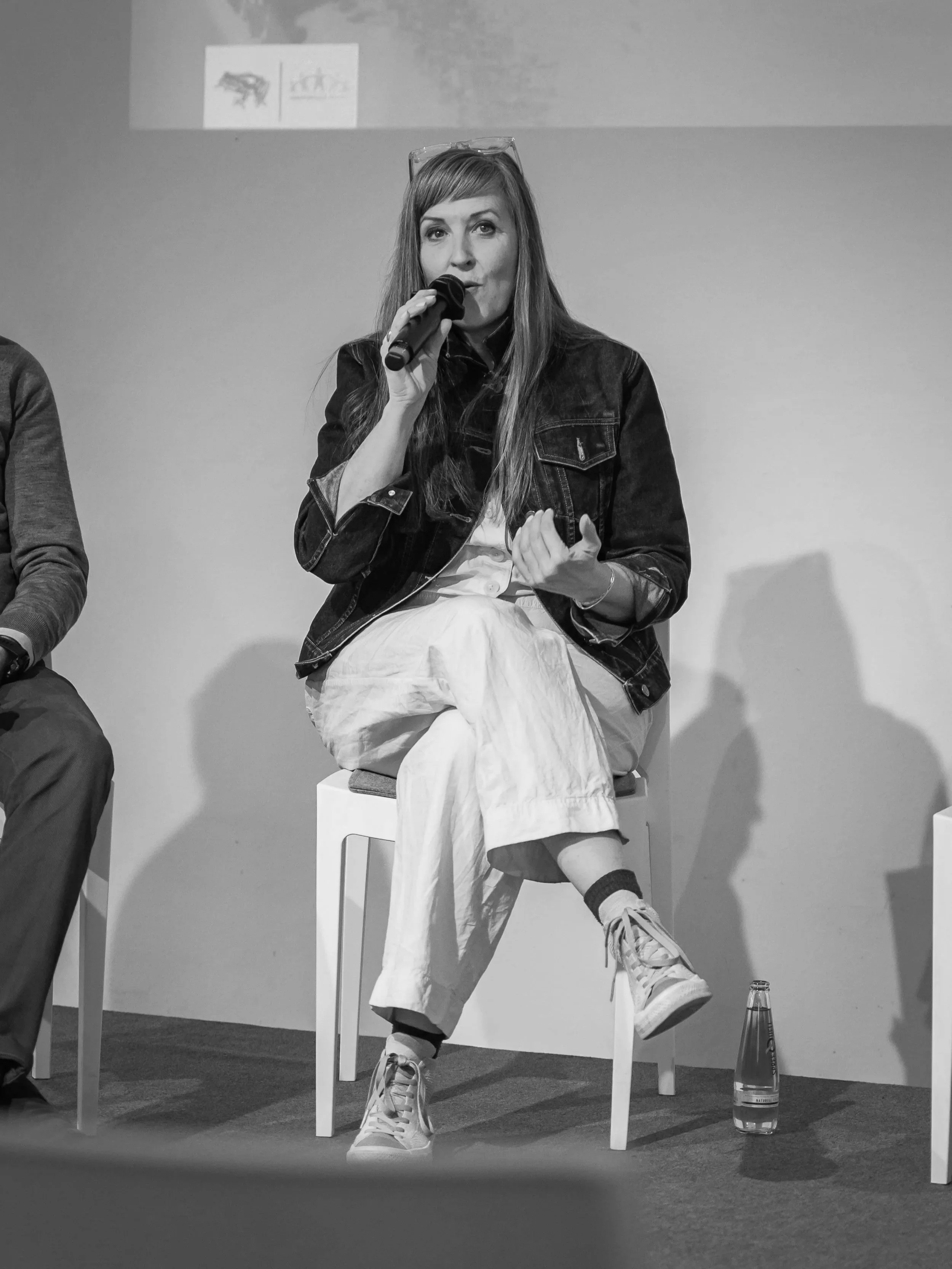 A woman with long hair, wearing a denim jacket, white pants, and sneakers, sitting on a stool and speaking into a microphone during a panel discussion. There are water bottles on the floor beside her and a plain wall in the background.