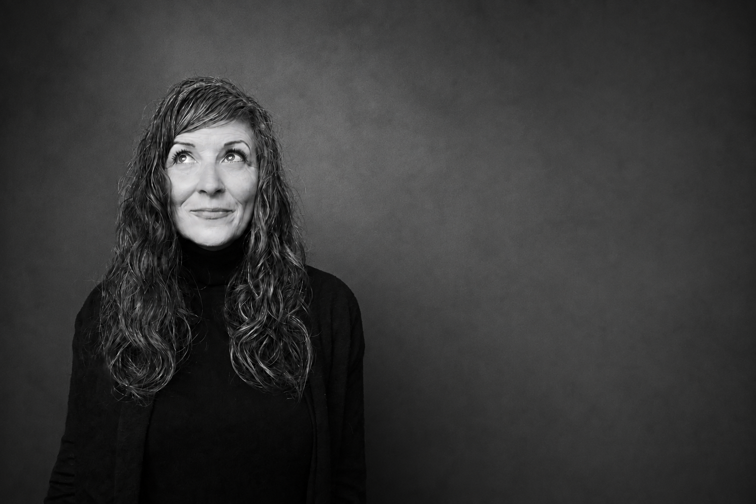 Black and white photo of a woman with long, curly hair looking upward with an amused expression, wearing a black turtleneck and a blazer, standing against a textured dark grey background.
