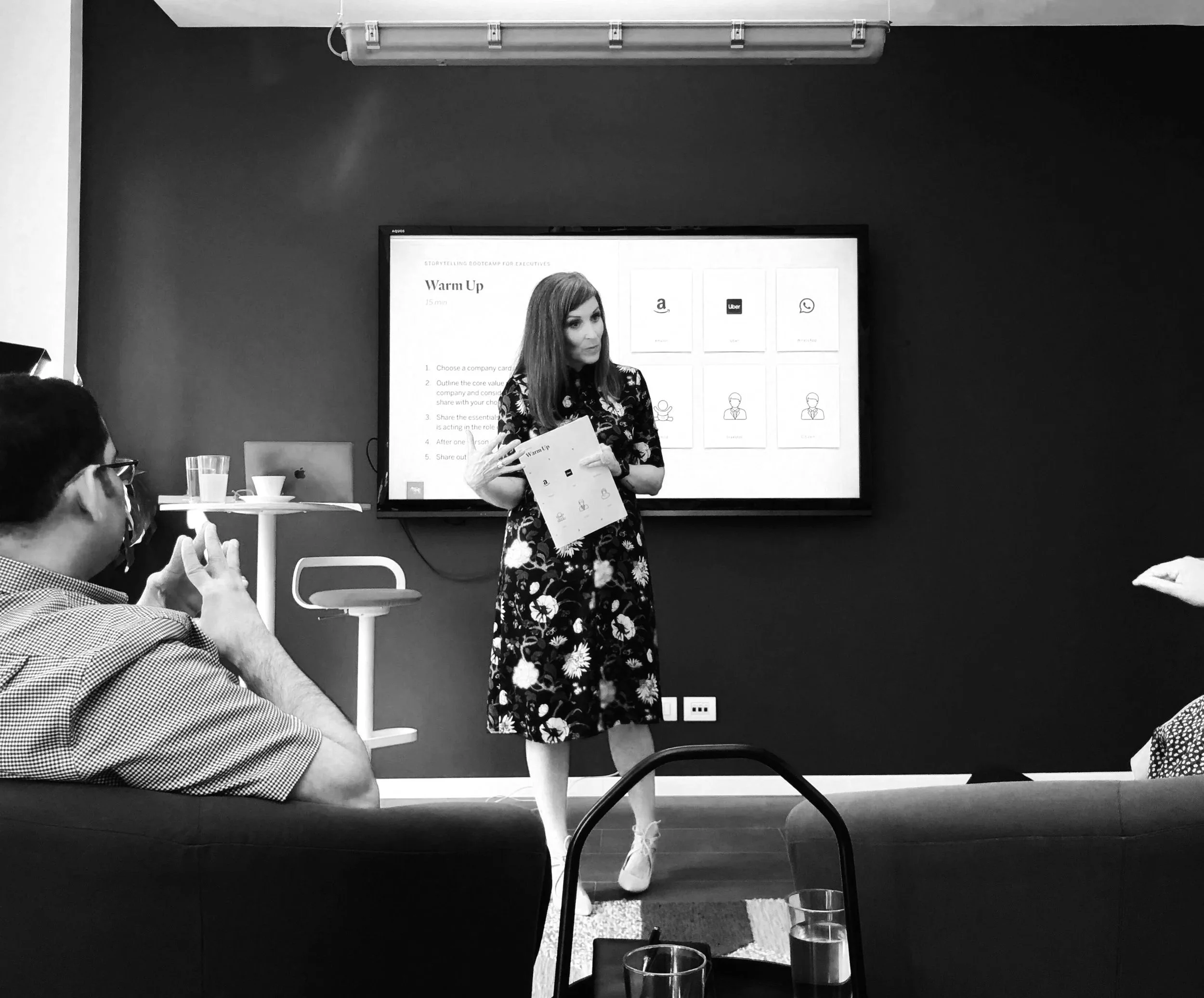 A woman standing and speaking in front of a large presentation screen in a conference room, with people seated and watching.