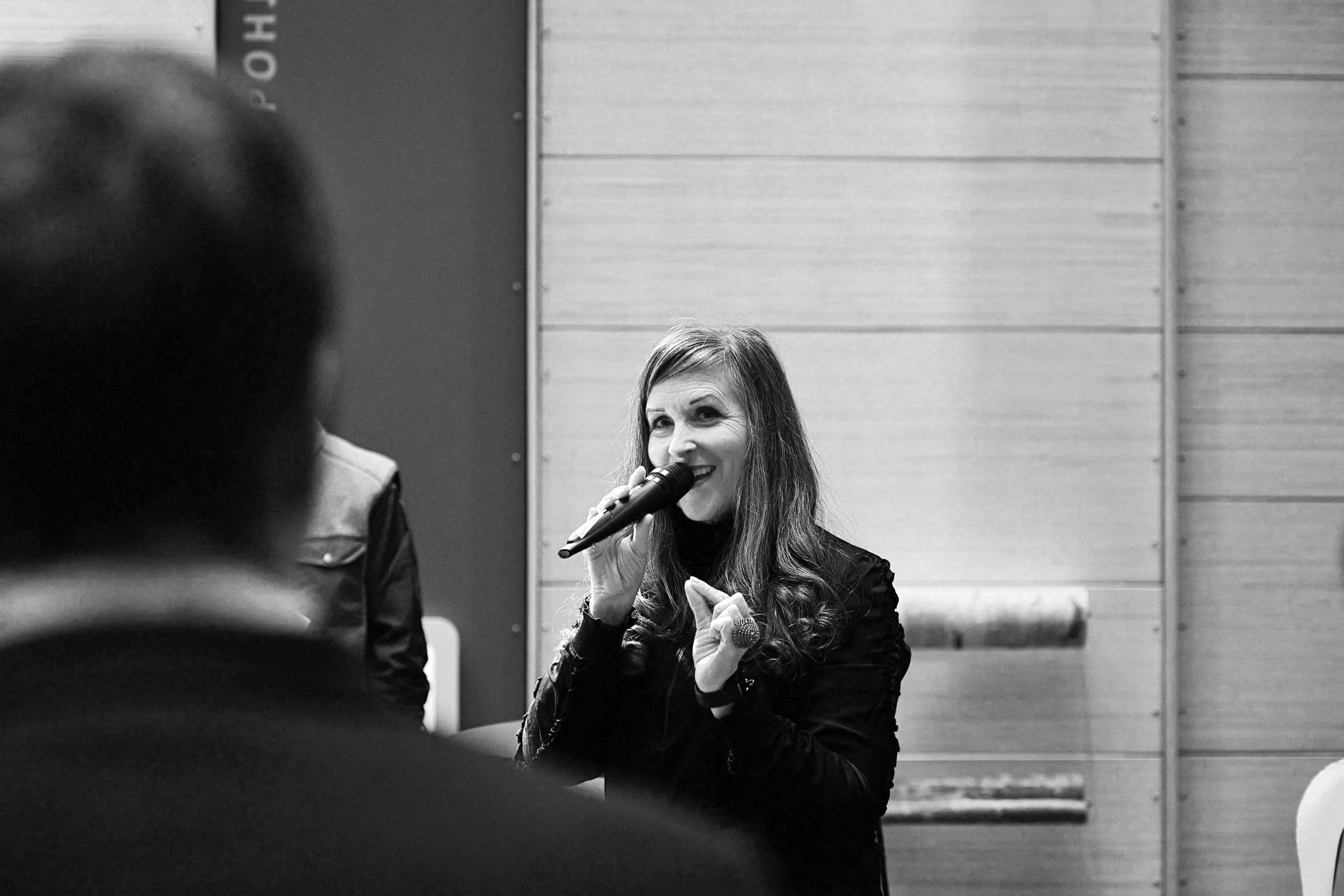 Woman with long hair holding a microphone and smiling, sitting in a room with wood-paneled walls, seen through the back of a person's head in the foreground.