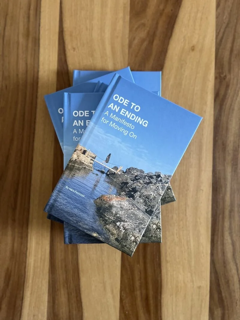 Several copies of a book titled "Ode to an Ending: A Manifesto for Moving On" by Kara Racheal, with a cover image of rocks and a person walking on a bridge over water, placed on a wooden surface.
