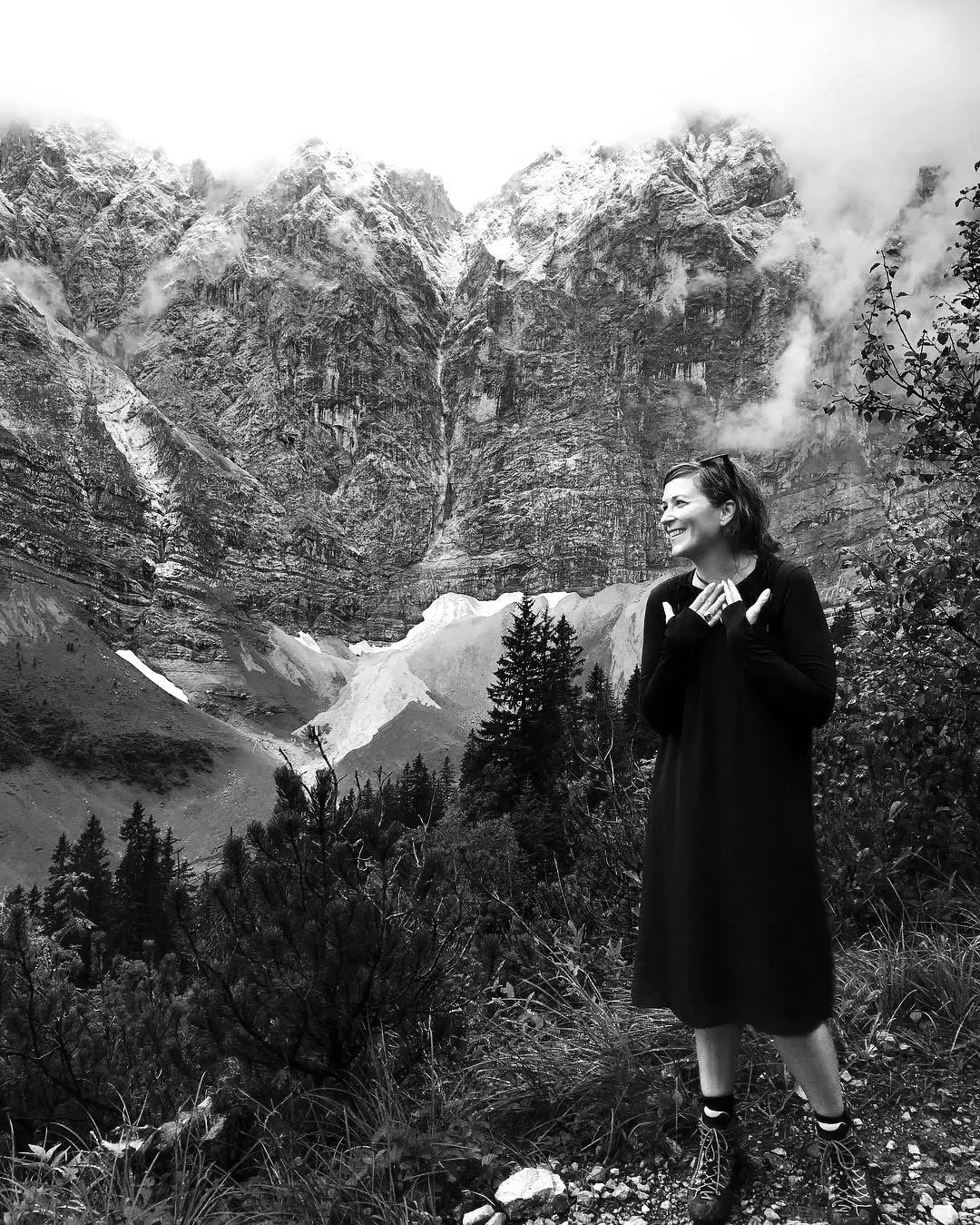 A woman standing outdoors in a mountainous area with snowy peaks, trees, and rocky terrain. She is smiling and wearing a long black dress with hiking boots.