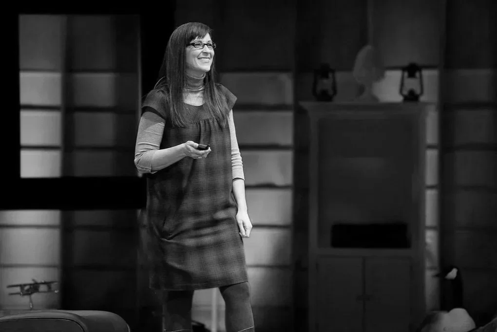 A woman with glasses and long straight hair stands smiling on stage, holding a remote control in her right hand, with wooden paneling background.