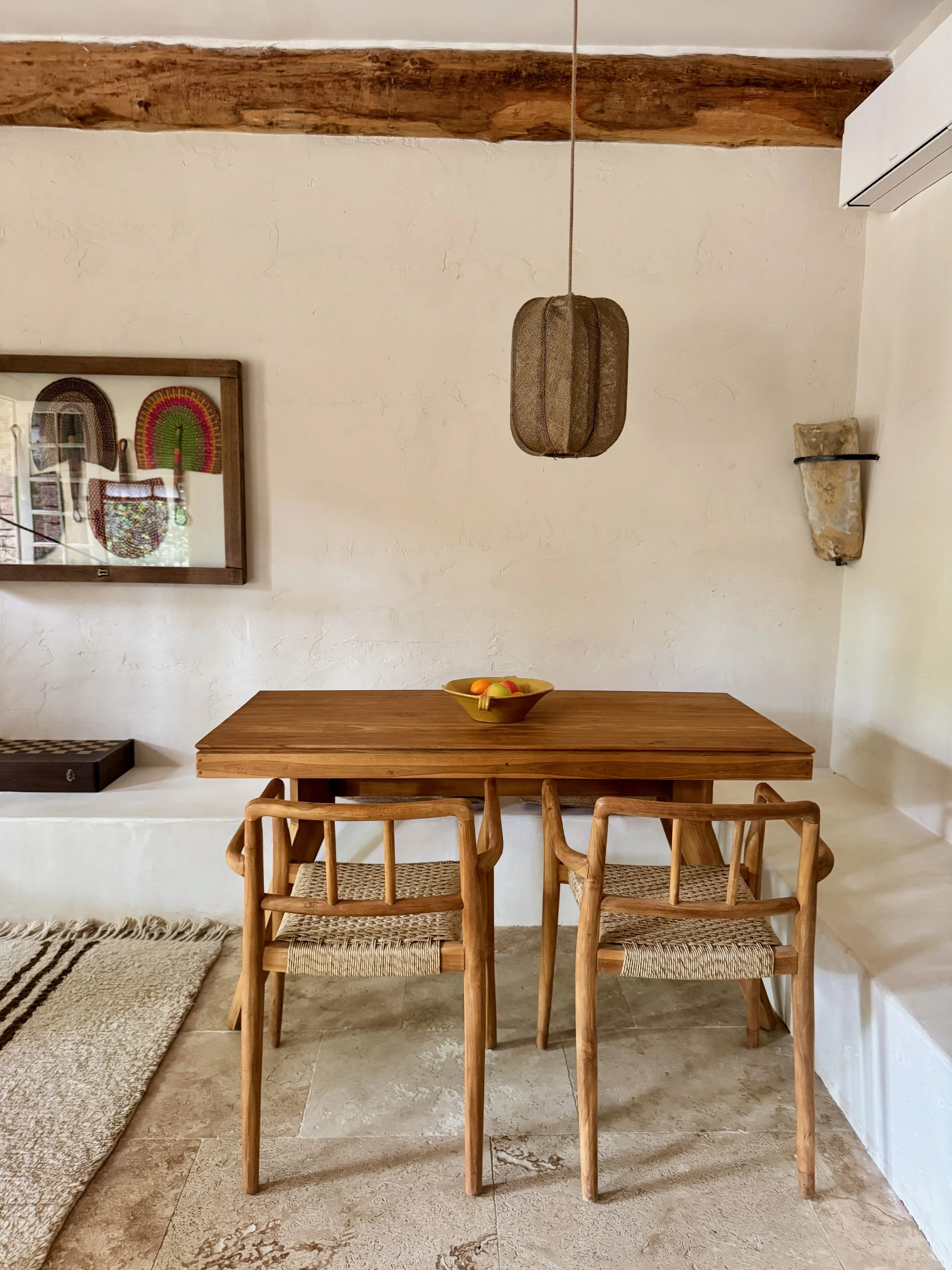 Une pièce avec une table en bois et quatre chaises en bois avec assise en corde. Sur la table, un bol de fruits. Au mur, un tableau avec des objets décoratifs, un plafond en bois et un éclairage suspendu en tissu. Mur blanc et sol en pierre.