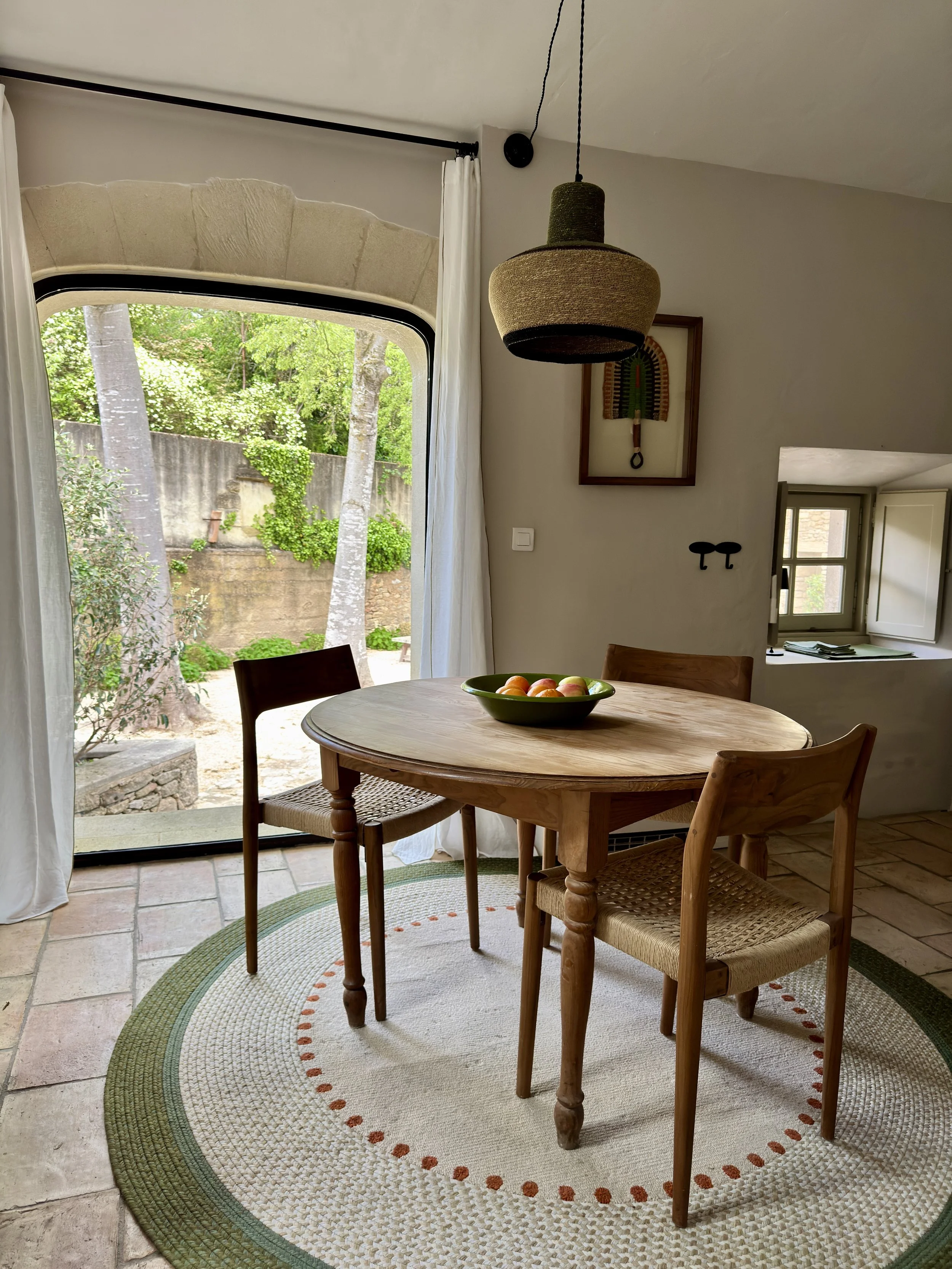 Une pièce lumineuse avec une table en bois ronde, entourée de quatre chaises en bois tressé. Un bol de pommes se trouve sur la table. Il y a une grande fenêtre donnant sur un jardin avec des arbres et une clôture. Une serrure et un tableau d'art colo